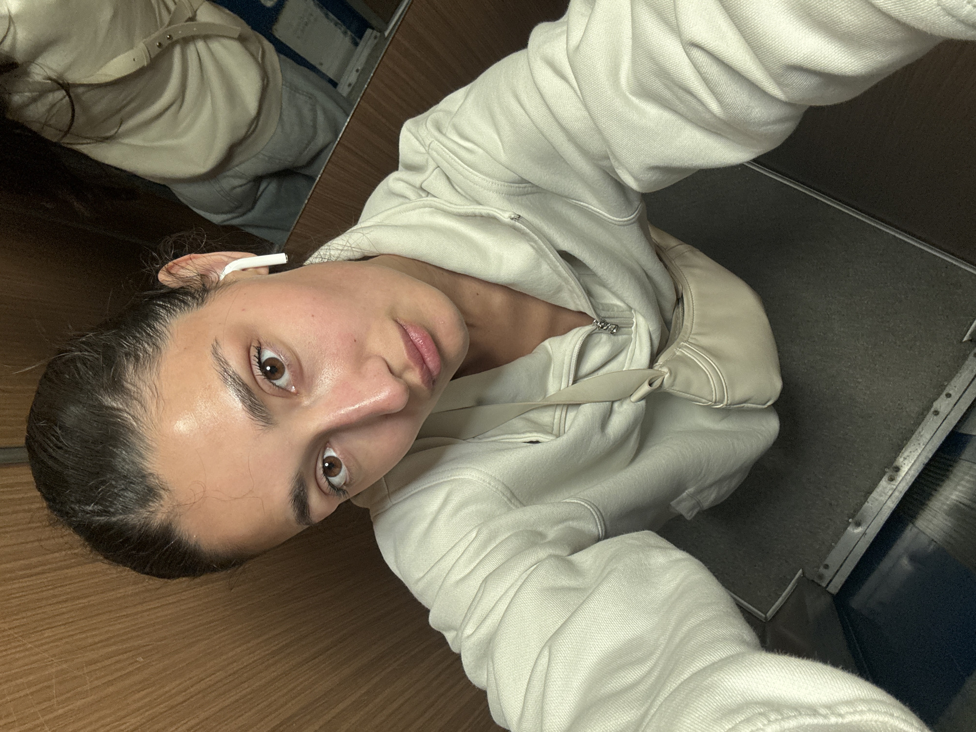 A young woman is captured in a selfie taken at an oblique angle, looking directly at the camera with a neutral to slightly pensive expression. She has prominent dark eyebrows and light brown eyes. Her dark hair appears pulled back, and she wears a white earbud in her visible left ear. She is dressed in a light-colored, possibly cream or off-white, hooded sweatshirt, which is partially unzipped. A matching or similarly colored shoulder bag or fanny pack is visible over her right shoulder.
The background elements suggest an enclosed space, likely the interior of a public transport vehicle such as a train or bus, consistent with travel in Bar, Montenegro. To her left, a warm-toned wooden paneling forms part of the wall. To her right, a dark grey, possibly carpeted or textured, floor is visible, extending towards a metallic strip and a dark blue fixture, which could be part of a seat base or interior paneling. A reflection in a mirror or window shows a blurred glimpse of her back and part of her clothing, along with other elements of the cabin interior. The lighting is artificial and somewhat subdued. There is no visible text in the image.