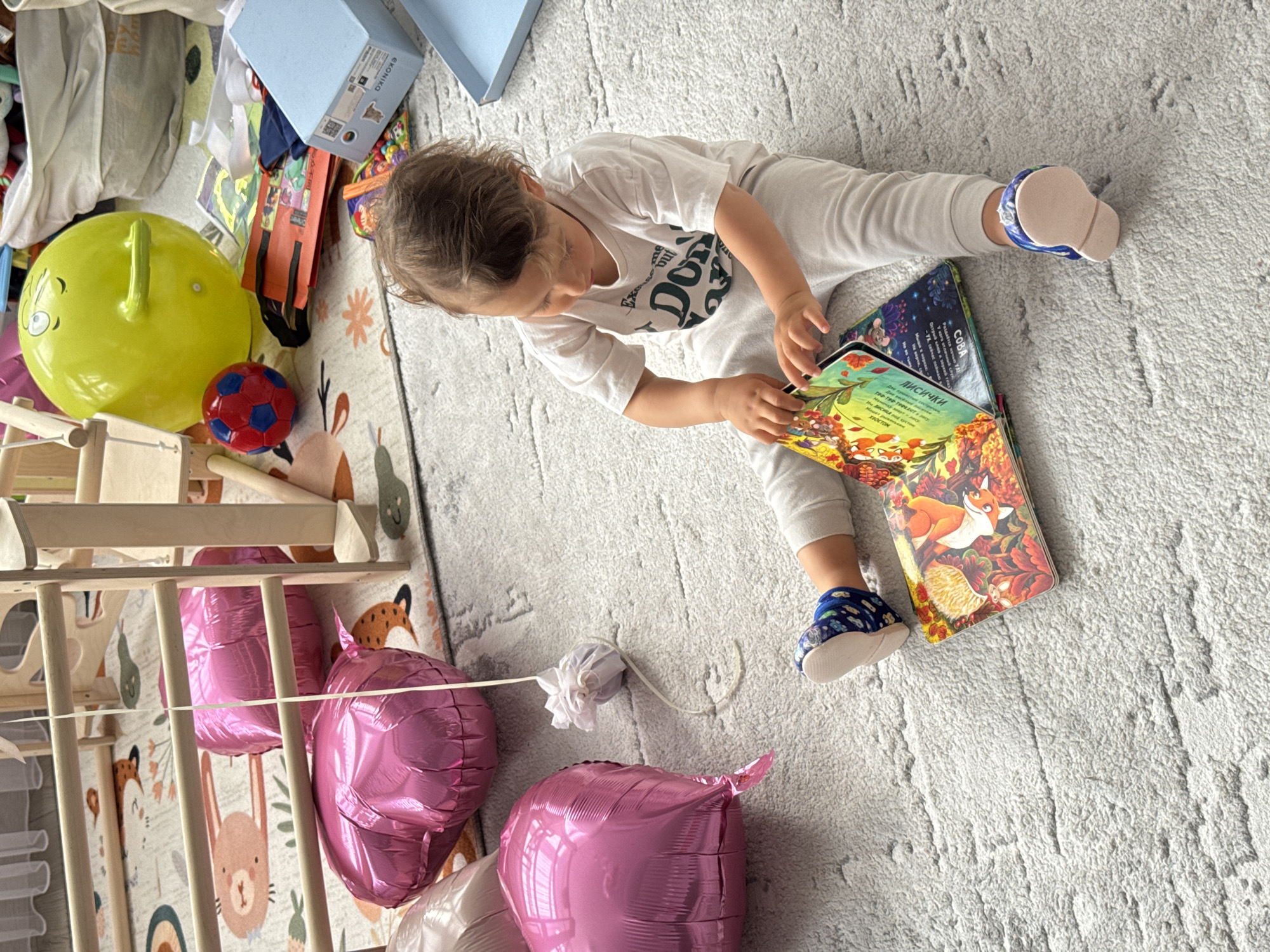 A child is sitting on a light gray carpet, reading a book with illustrations of a fox and other animals. The child is wearing a white shirt, light-colored pants, and blue and white shoes. The book is open to a page with text in Russian.
Around the child, there are several toys, including a yellow riding toy, a red and blue ball, pink balloons, and a wooden structure that appears to be a climbing frame. There are also blue boxes and other items scattered around the area. The background shows a rug with a fox pattern. The scene appears to be inside a home. It is not possible to confirm the city or country from the provided image.