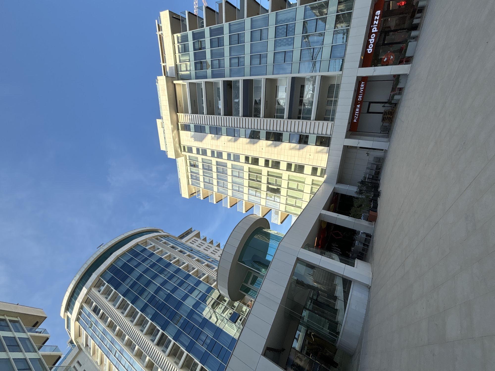 This is a low-angle, wide shot of a modern, multi-story building complex situated in Budva, Montenegro. The complex features sleek architecture with extensive use of glass and light-colored materials. Two prominent buildings dominate the frame, one with a curved facade of blue glass and balconies, and another taller, more rectangular building with a grid of windows and balconies. The ground level of the complex is characterized by a spacious, paved outdoor area and covered walkways leading to various businesses. Visible signage indicates a "PIZZERIA - DELIVERY" establishment and a "dodo pizza" restaurant. The sky above is a clear, bright blue. The scene depicts an urban environment with commercial and residential spaces, bathed in sunlight.