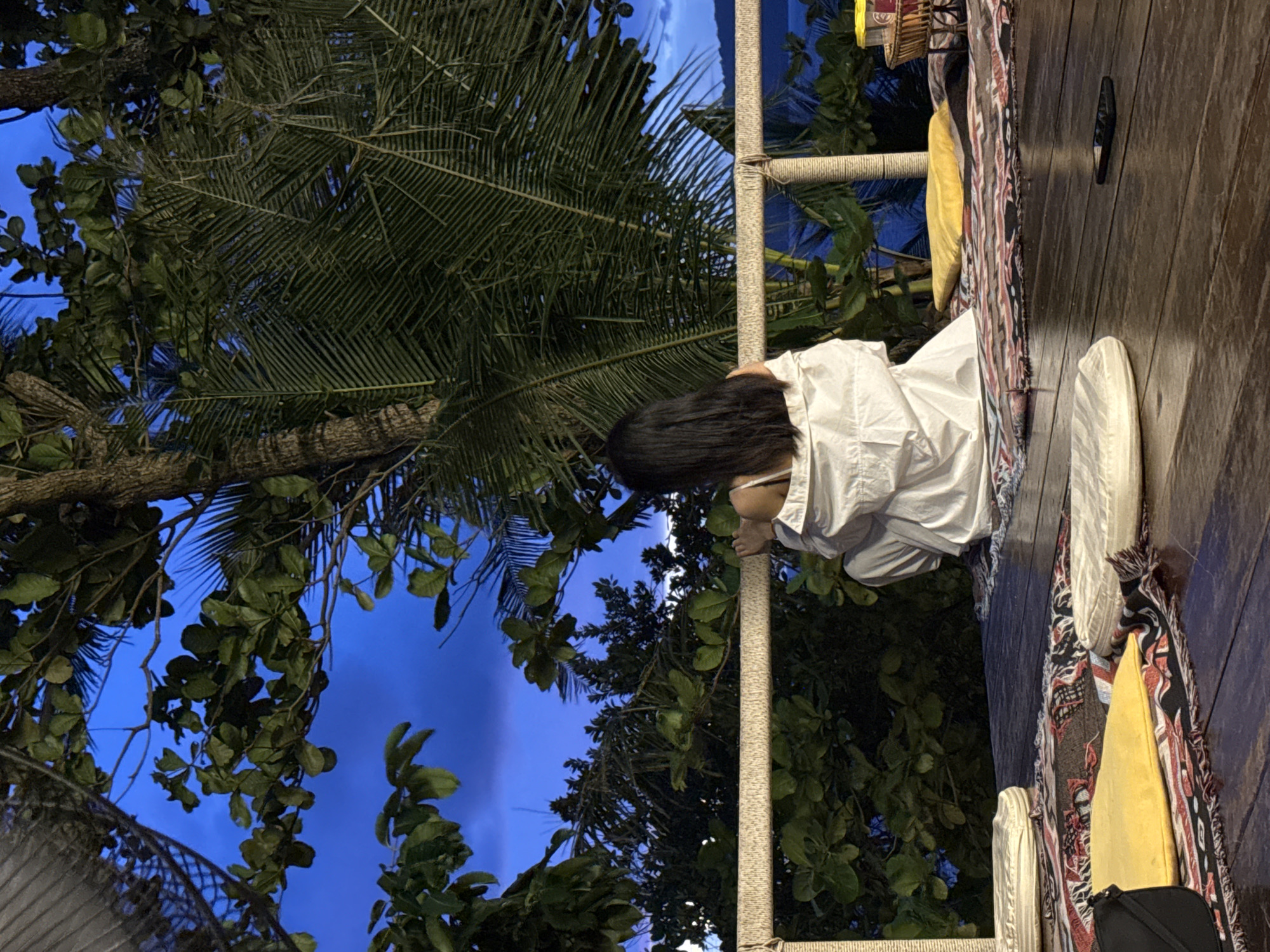 A person wearing a white shirt is kneeling on a wooden deck. They are facing away from the camera, with their head bowed and long, dark hair covering their face. The person is leaning against a thick, rope-wrapped railing.
The scene appears to be outdoors, with lush green foliage and palm fronds visible against a deep blue twilight sky. The lighting suggests it is either early morning or late evening. The wooden deck is adorned with colorful textiles, including a patterned blanket with red, black, and white designs, and a bright yellow cushion. White shoes are placed on the deck near the railing.
The overall atmosphere is peaceful and serene, with the natural surroundings creating a tranquil setting. There is no visible text in the image.
