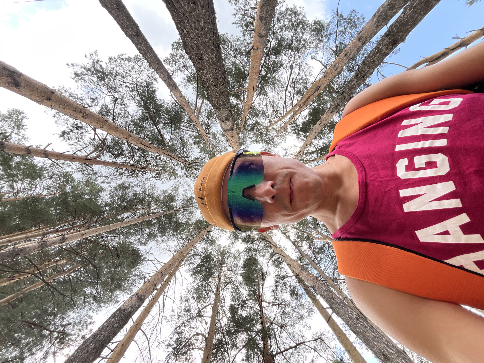 In a low-angle shot from within a pine forest, a person wearing a bright orange and pink athletic top, a tan cap, and reflective sunglasses is positioned in the foreground. The person's face is partially visible, looking upwards towards the camera. The text "CHANGING" is visible in large white letters on the pink part of the shirt. The background consists of the canopy of numerous tall, slender pine trees reaching towards a bright, cloudy sky. The scene suggests an outdoor activity, possibly running or cycling, given the athletic attire. The specific location in Chaikovsky, Russia, is not discernible from the visual information provided, but the presence of pine trees suggests a natural, wooded environment.