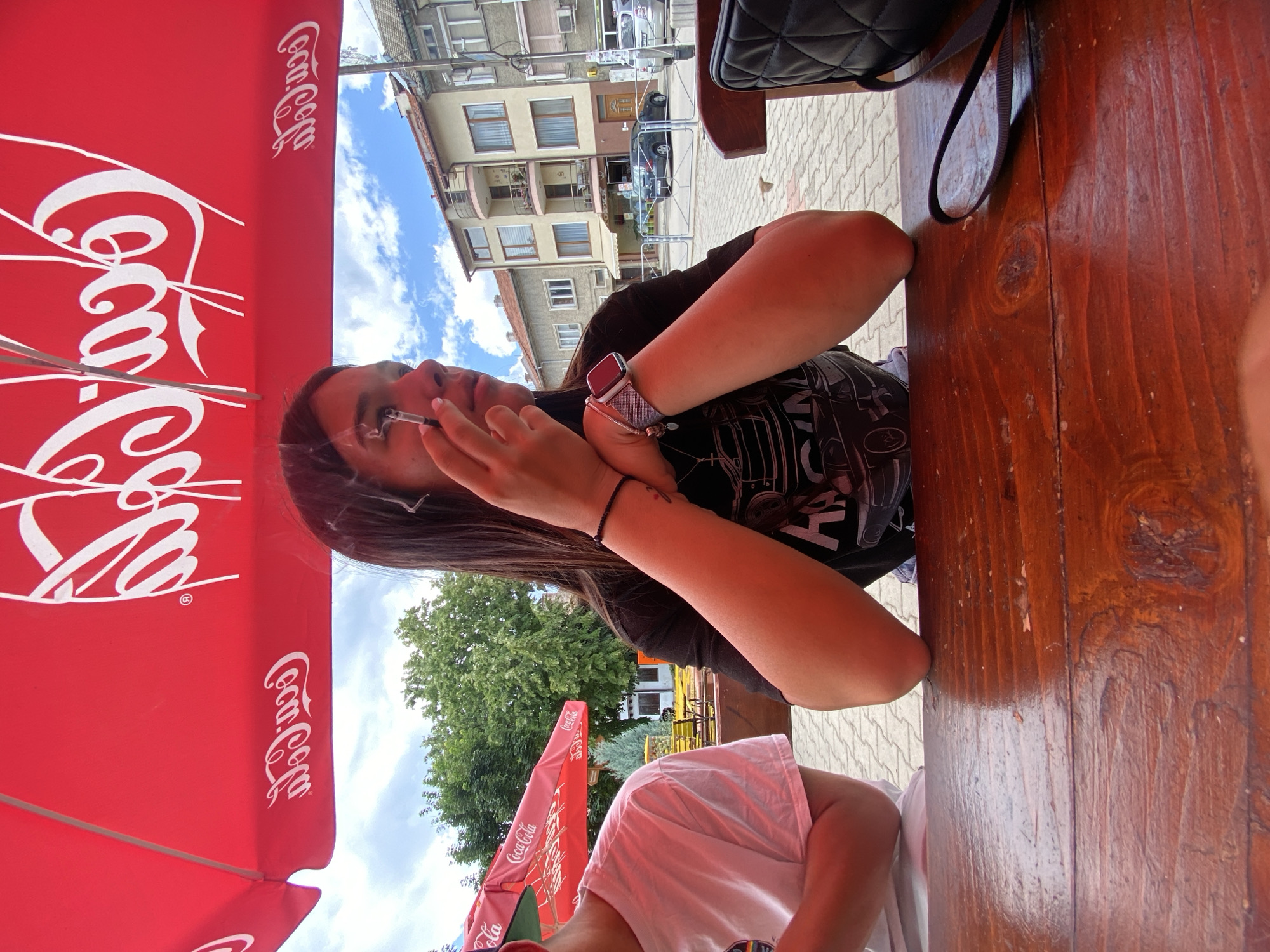 The image shows two individuals sitting at a wooden table outdoors under a red Coca-Cola umbrella. A woman on the right is applying makeup to her eye with a mascara wand while holding a cigarette in her other hand. She is wearing a black t-shirt and a smartwatch on her left wrist. To her left, a person wearing a white t-shirt is partially visible. In the background, there are buildings, trees, and a cloudy blue sky. The scene appears to be a street-side cafe or restaurant in Sevlievo, Bulgaria.
