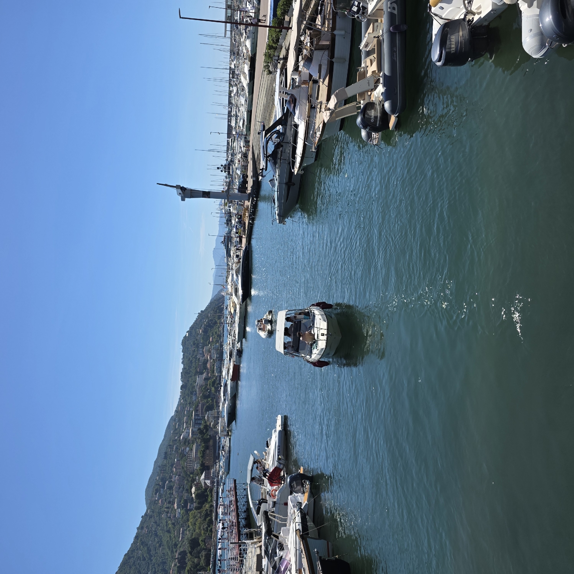 This is a high-angle, medium shot of a marina in Rapallo, Italy, on a clear, sunny day. The foreground is dominated by dark blue water with ripples and reflections of sunlight. Several boats are moored in the marina. In the middle ground, a small white motorboat with a canopy is moving away from the viewer. It carries at least four people, two of whom appear to be standing. To the left, several other motorboats are docked, including one with a prominent black Yamaha outboard motor.
In the background, a long pier with railings and a red walkway extends into the water. Numerous sailboats are docked beyond the pier, their masts creating a dense forest of vertical lines against the sky. A lighthouse-like structure with a beacon is visible on the far right of the pier. Beyond the marina, the hills of Rapallo rise steeply, covered in lush green trees and dotted with buildings, indicating a coastal town nestled against a mountainous backdrop. The sky is a vibrant, cloudless blue.