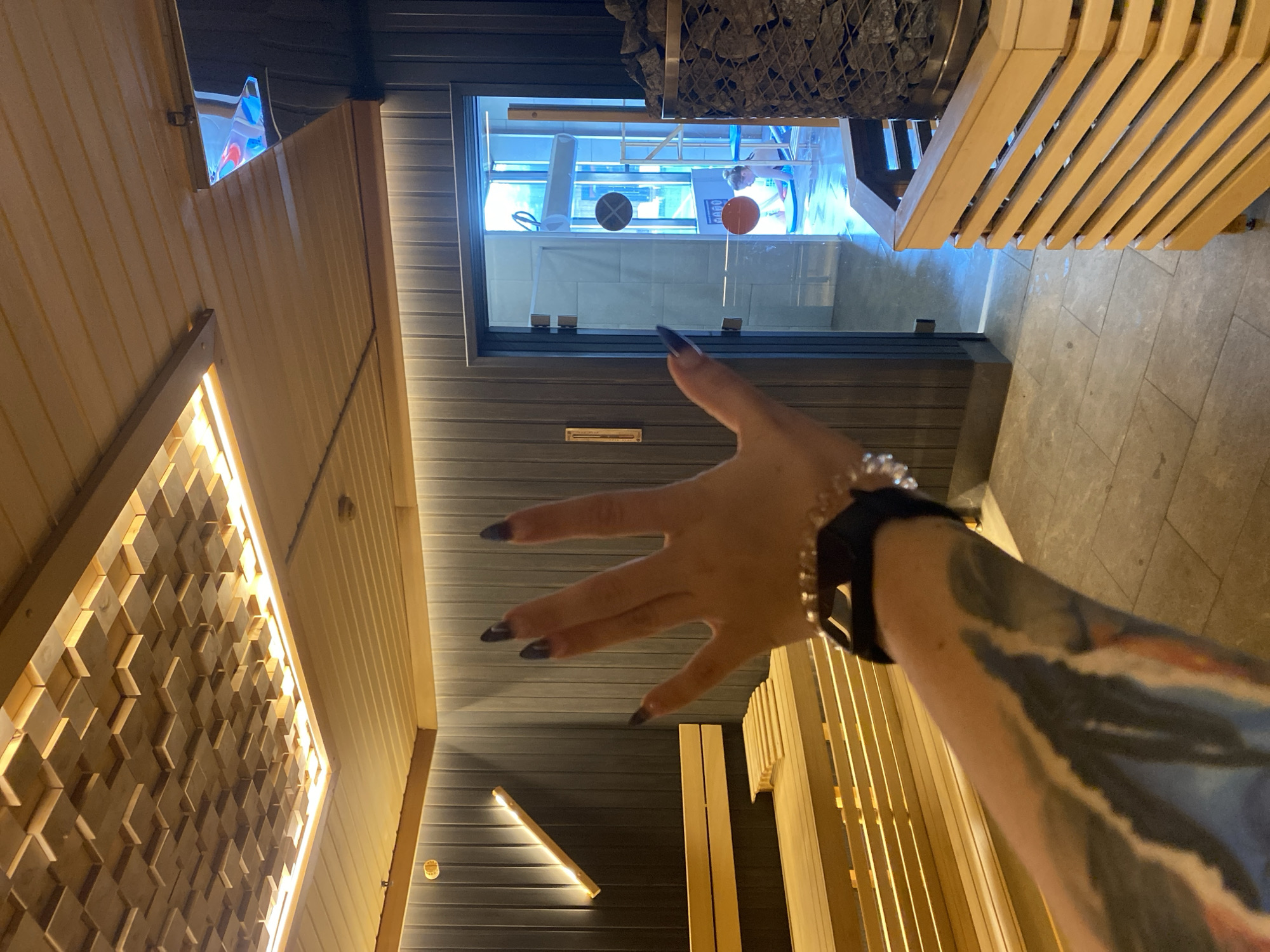 The media file shows a first-person perspective of a person in a modern sauna. The person's tattooed arm and hand, with dark nail polish, are prominently in the foreground, reaching out towards the camera. The hand is open, with fingers spread. A beaded bracelet is visible on the wrist.
The sauna is constructed with light-colored wood paneling on the walls and ceiling, with some sections featuring a geometric wooden inlay illuminated by hidden lights. A glass door or window leads to another area, which appears to be a spa or pool facility, evidenced by the bright blue lighting, tiled walls, and some equipment visible through the glass. A sauna heater filled with stones is situated in the upper corner of the frame, above the glass door. Wooden benches are visible on the right side of the frame. The flooring appears to be tiled.
The lighting suggests it might be evening or an indoor environment. The overall impression is one of relaxation and modern spa amenities. No specific time of day or weather conditions can be determined from the image itself. No visible text is present in the image. The location context of Votkinsk, Russia is provided externally and is not directly inferable from the visual content alone.