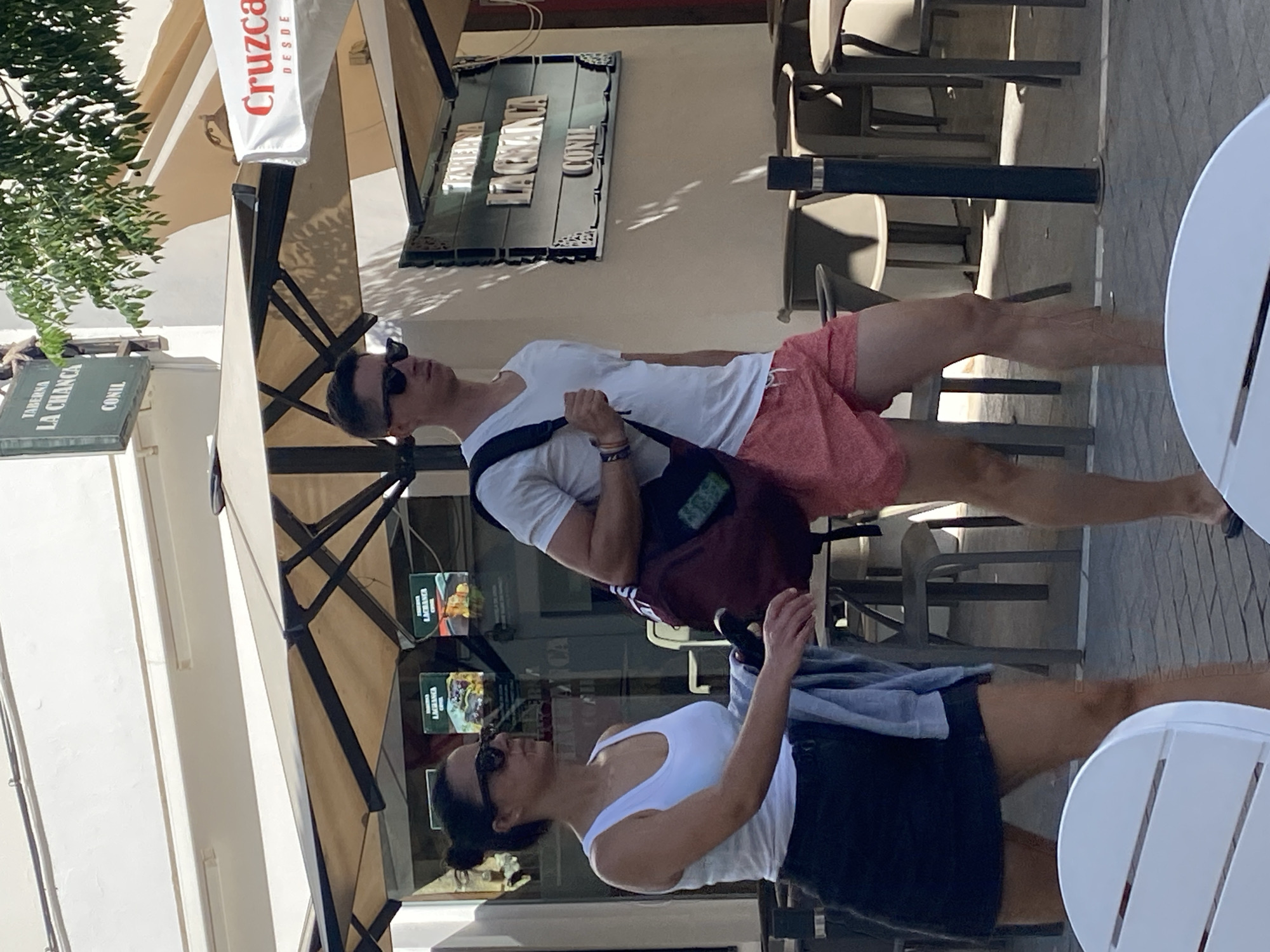 Two people, a woman and a man, are walking on a sidewalk in front of a building with an awning and umbrellas. The woman, on the left, is wearing sunglasses, a white tank top, and a dark skirt. She has her dark hair pulled back in a bun and is carrying a light-colored garment over her left arm. The man, on the right, is wearing sunglasses, a white t-shirt, and red patterned shorts. He has a dark bag slung over his right shoulder. Behind them, a sign for "TABERNA LA CHANCICA CONIL" is visible on the building. Tables and chairs for an outdoor seating area are set up in front of the establishment. The street is paved with cobblestones. The lighting suggests it is daytime, likely a sunny day given the shadows.