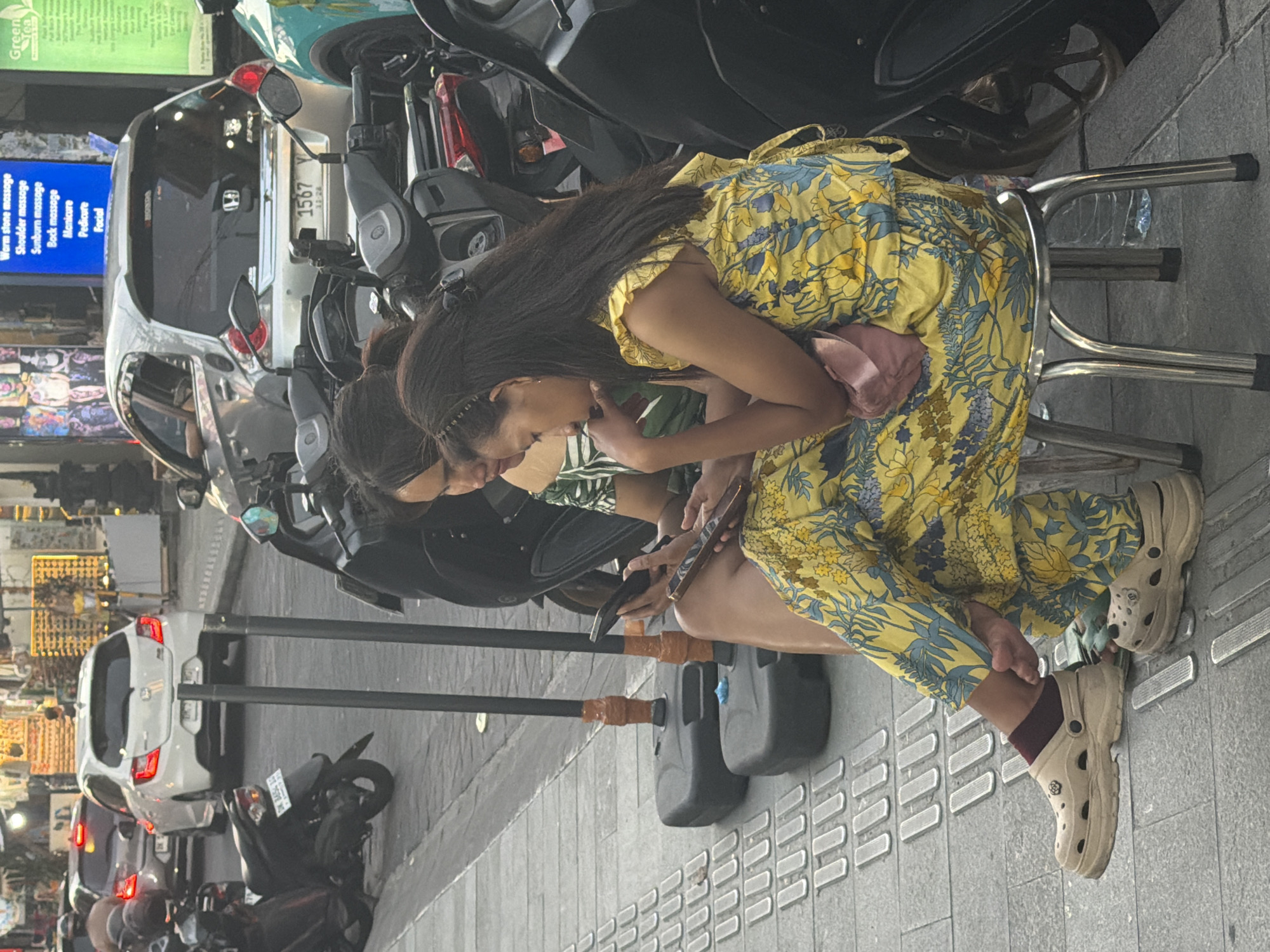 In Kuta, Indonesia, two women are seated on a metal stool on a sidewalk. The woman on the left wears a green and white striped top and is looking at her smartphone. The woman on the right wears a yellow dress with a floral pattern and is also looking at her smartphone. Both women are wearing clogs. Behind them, motorcycles are parked along the sidewalk. In the background, cars are visible on the street, and shops line the street.