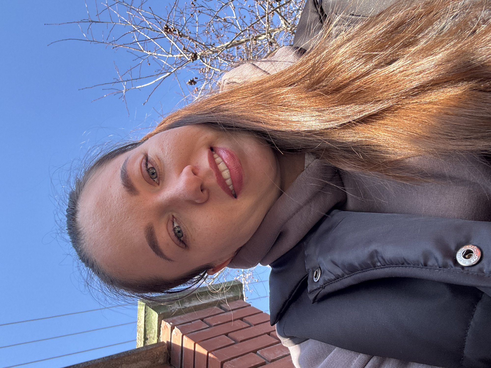 A female individual is depicted in a low-angle selfie, positioned outdoors in Ферма, Russia. She has long, reddish-brown hair, light-colored eyes, and a light complexion, exhibiting a closed-mouth smile. The individual is dressed in a dark, possibly black, puffer vest over a light-colored hooded top, with a coordinating scarf visible around her neck. The background features a clear blue sky. In the upper left quadrant, a reddish-brown brick wall with a concrete capping is partially visible, with multiple parallel utility lines traversing the sky above it. To the upper right, the bare branches of a deciduous tree, bearing dark seed pods, are visible against the sky. The scene is illuminated by direct sunlight.