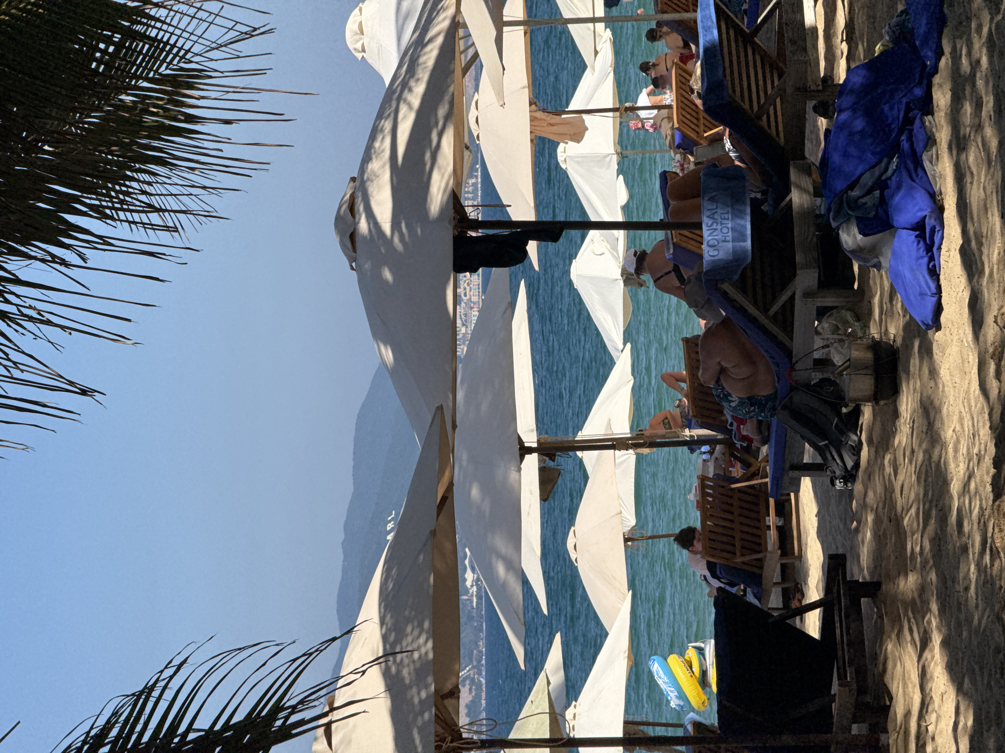 This image depicts a sunlit beach scene in Nha Trang, Vietnam. The foreground consists of sandy terrain with distinct shadows cast by unseen overhead palm fronds, visible as dark silhouettes in the upper left and right corners. Numerous light-colored, round beach umbrellas are open and distributed across the sand, providing shade.
Under these umbrellas and in direct sunlight, multiple individuals are present. Several people are seated or reclining on wooden and blue fabric lounge chairs. One individual, wearing a white cap and dark top, is seated facing the ocean slightly to the left of center. Another person is lying face down on a wooden lounge chair in the mid-left. A blue towel marked "GONSALA HOTEL" is draped over a blue lounge chair in the central foreground. Various items of clothing are visible, including a dark garment hanging from an umbrella pole and blue towels piled on the sand in the lower right foreground. Near the water's edge on the left, a blue and yellow inflatable ring is present.
The mid-ground features the blue-green ocean with minimal surface agitation. The background reveals distant mountains and hills under a clear, bright blue sky. A faint urban skyline is visible along the distant coastline, and the letters "MRL" are discernable on a prominent hillside. All visible individuals appear to be in a state of leisure, typical of a beach environment.