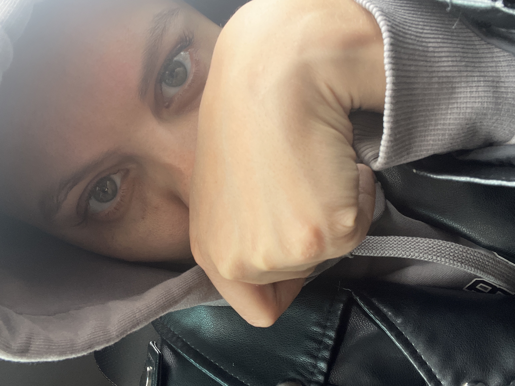 This close-up, angled self-portrait captures a person's upper face and clothing. The subject's light-colored eyes, likely green or hazel, look directly at the camera, with visible reflections indicating a light source. Their skin appears fair. A hand is positioned near the face, partially obscuring the lower part of the face, suggesting a shy, intimate, or contemplative pose.
The person is wearing a light gray hooded sweatshirt (hoodie) with a ribbed cuff visible on the sleeve. The hood is partly pulled up around the head. Over the hoodie, they wear a black jacket, appearing to be made of leather or faux leather, with visible stitching and possibly metal hardware detail. The lighting is soft and consistent, suggesting an indoor or sheltered daytime setting, though no specific background elements are discernible within the frame. The overall atmosphere feels personal and slightly reserved.
In one of the cropped hints, a drawstring from the hoodie is visible, and on the gray fabric of the hoodie itself (perhaps an interior tag or detail), there is a faint, indistinct white rectangle with dark markings, which could be part of a logo or text but is unreadable. The location context provided for this image is Chaikovsky, Russia, though no specific visual cues for this location are present in the image itself.