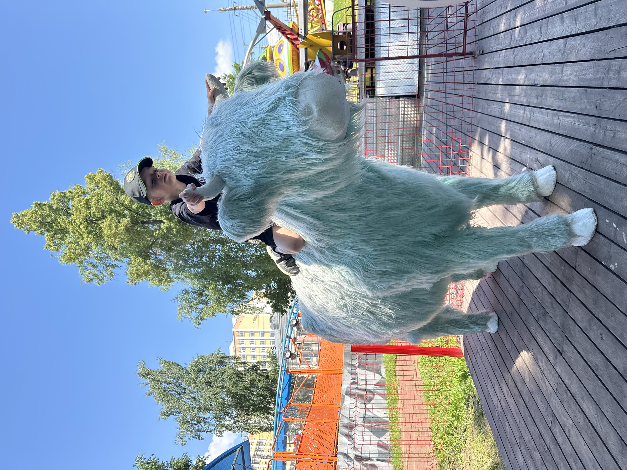 A young boy wearing a black t-shirt and a baseball cap rides on the back of a large, fluffy, light blue cow sculpture. The cow is positioned on a wooden platform in an outdoor amusement park or playground. In the background, there are several trees, a bright blue sky with scattered clouds, and buildings that appear to be in a city. There are also parts of amusement park rides visible, including an orange railing and a yellow and red ride with a spherical component. The scene is set during the daytime, under clear, sunny skies. This image was taken in Izhevsk, Russia.