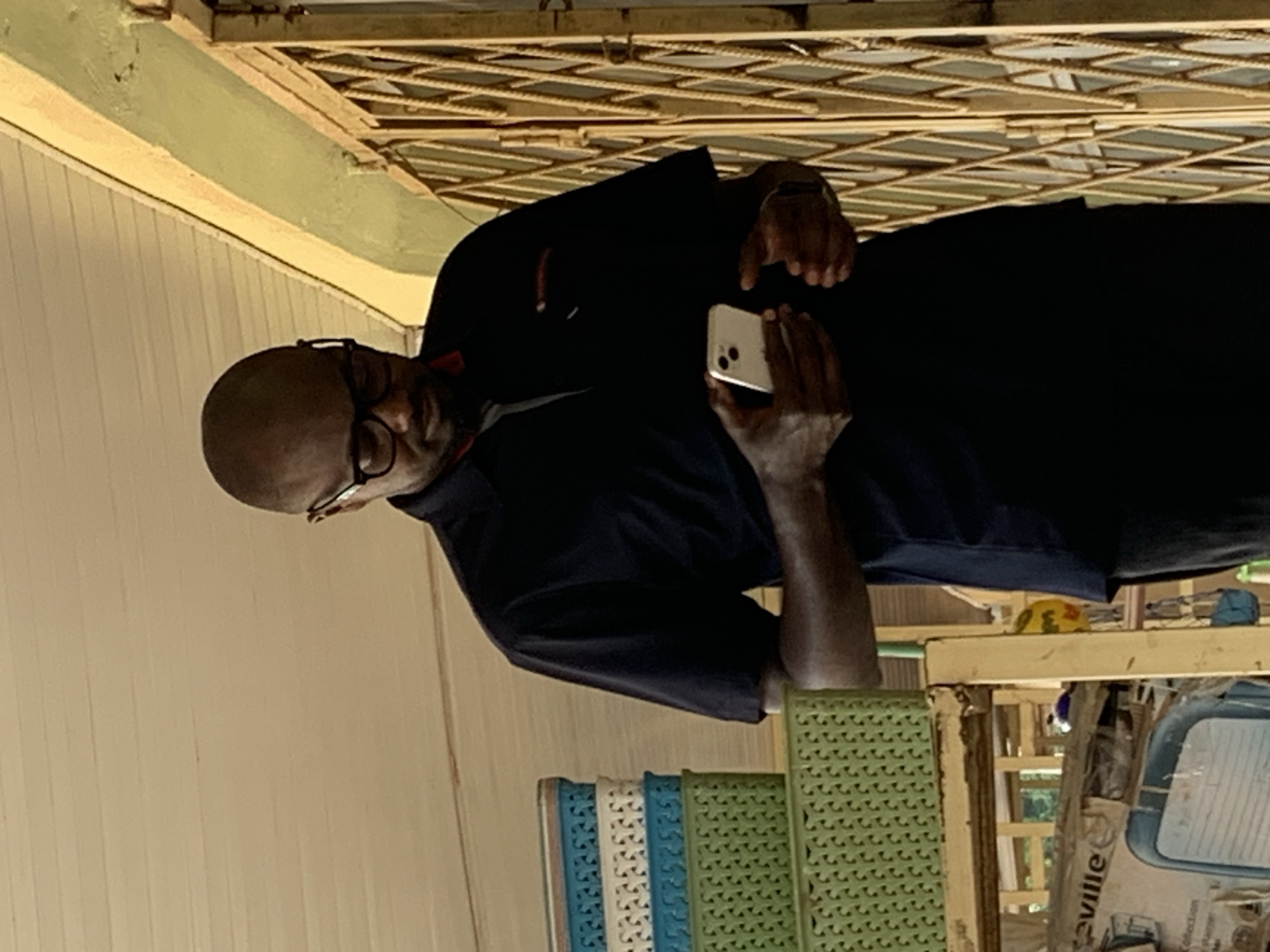 A bald Black man wearing glasses and a dark blue polo shirt with a red collar stands outdoors, looking down at a white smartphone in his hands. He is positioned in front of a building with a yellow exterior wall and a latticed metal gate. To his left, there is a stack of colorful plastic baskets, including blue, white, and green ones. The man's left hand holds the phone while his right hand is near the bottom of the phone. The environment suggests a commercial or storage area in Jos, Nigeria.