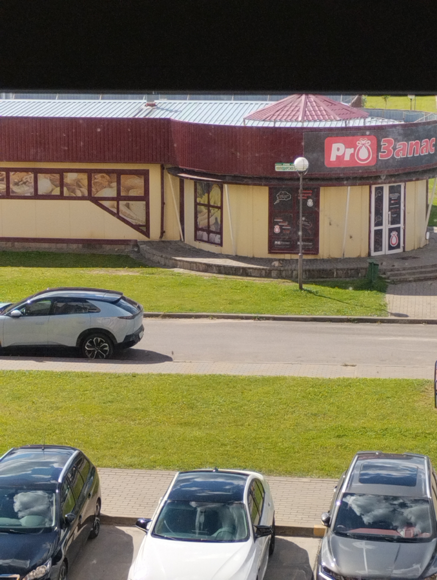 The image shows a street-level view looking down at a building with a curved facade and a red, conical roof section. The building appears to be a commercial establishment, possibly a bakery or cafe, with a prominent sign that reads "Pro Banac" with a red circular logo containing a white abstract shape. The building has large windows and doors, some of which are decorated with posters or graphics. In front of the building, a light-colored SUV is parked on the side of the road. Further down, three cars are parked in a lot: a black SUV, a white car, and a silver SUV. The scene is set in a well-lit, outdoor environment with green grassy areas and paved surfaces. The overall impression is one of a typical urban setting in Astravyets, Belarus, on a sunny day.