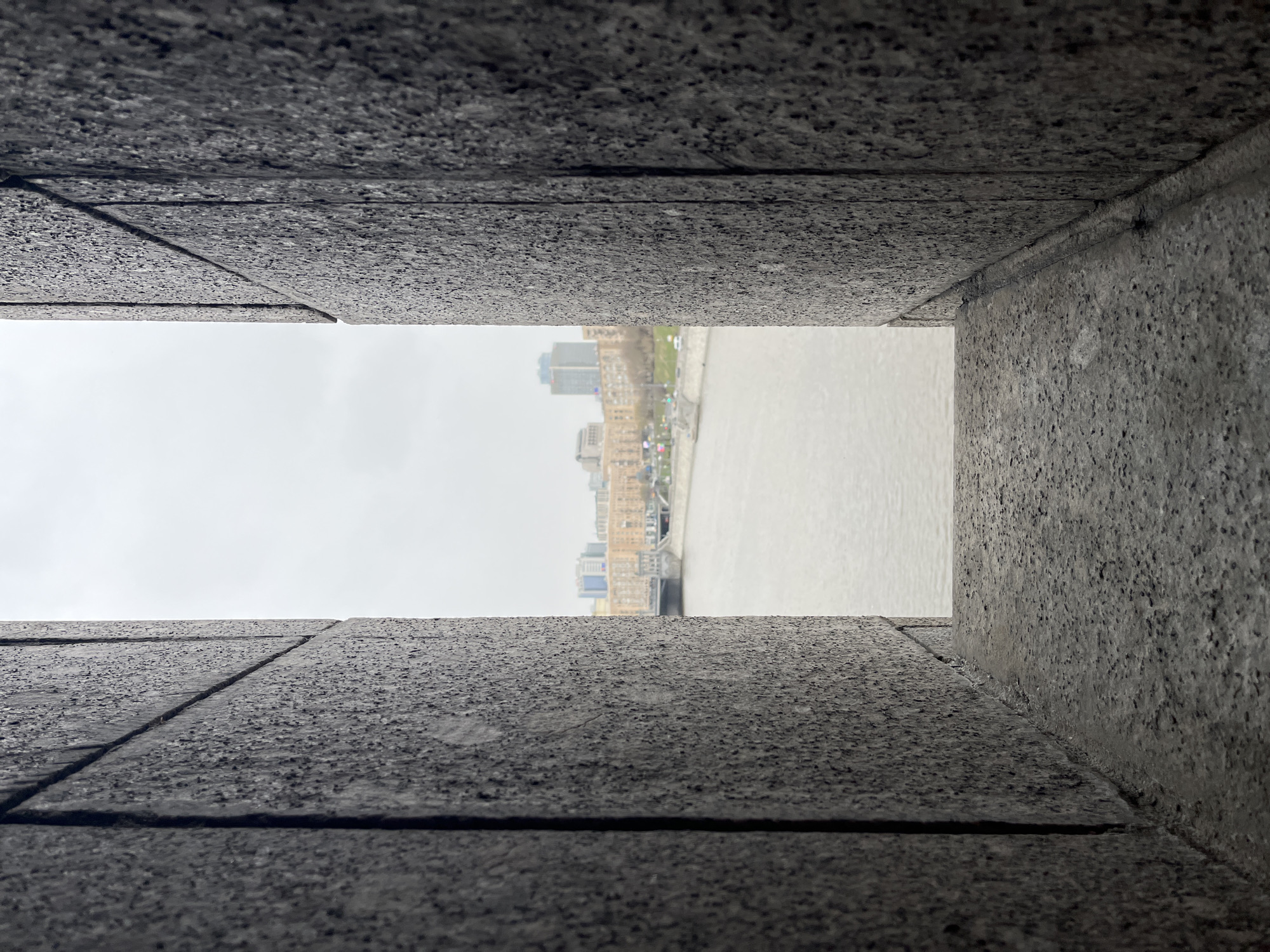 The image depicts a view of Moscow, Russia, seen through a narrow opening in a gray, textured stone structure. The opening is rectangular and framed by stone blocks. In the distance, a river flows horizontally across the frame. The riverbanks are lined with buildings, including some taller structures. The sky above the buildings and river is overcast. The stone structure appears to be a part of a larger architectural element. The angle suggests that the image was taken from a low perspective, looking upwards and outwards through the opening.