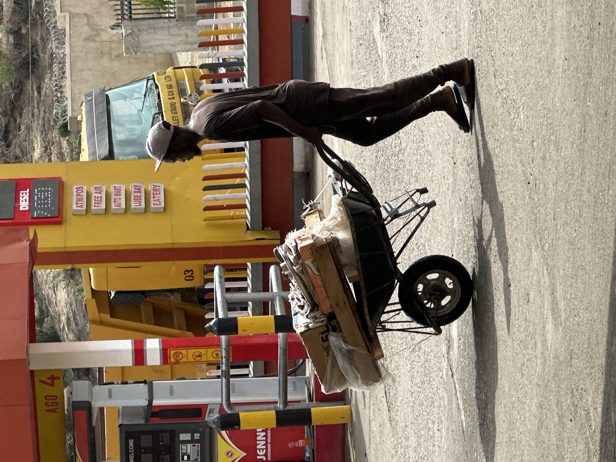 The image depicts a person pushing a heavily loaded wheelbarrow at a gas station in Gura Top, Nigeria, under bright, sunny conditions. The individual, seen from behind and to the side, wears a dark t-shirt, dark trousers, and a white baseball cap. They are actively pushing the wheelbarrow, which is filled with an assortment of items including cardboard boxes, wooden planks, and white plastic bags, across a paved area.
The setting is clearly a fuel station, characterized by prominent yellow and red structures, visible fuel pumps, and service signage. In the background, a large yellow truck is parked, bearing the text "LEY GYADO & CO NIG LTD". Beyond the station, a rugged, rocky hillside is visible, partially topped with a barbed wire fence.
The strong sunlight creates distinct shadows of the person and the wheelbarrow on the ground. Visible text on the gas station's signs includes "DIESEL" on a red price board, "AGO 4" on a pump's yellow stand, and a series of service indicators: "ATM/POS", "FREE AIR", "AUTO MART", "LUBE BAY", and "EATERY". Another sign near a pump displays "WELCOME" and "JENNY INVESTMENT NIGERIA L", while a yellow pillar is marked "03".