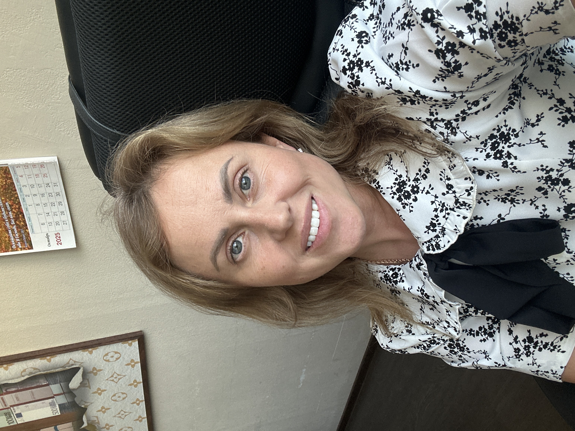 The image is a selfie of a woman with blonde hair and blue eyes, wearing a black and white floral print blouse with a black bow. She is sitting in an office chair in front of a wall with a framed picture and a calendar. The calendar shows October 2025. The framed picture appears to be a collage of European currency and a Louis Vuitton pattern. There is no discernible information in the image that places the location in Sochi, Russia.