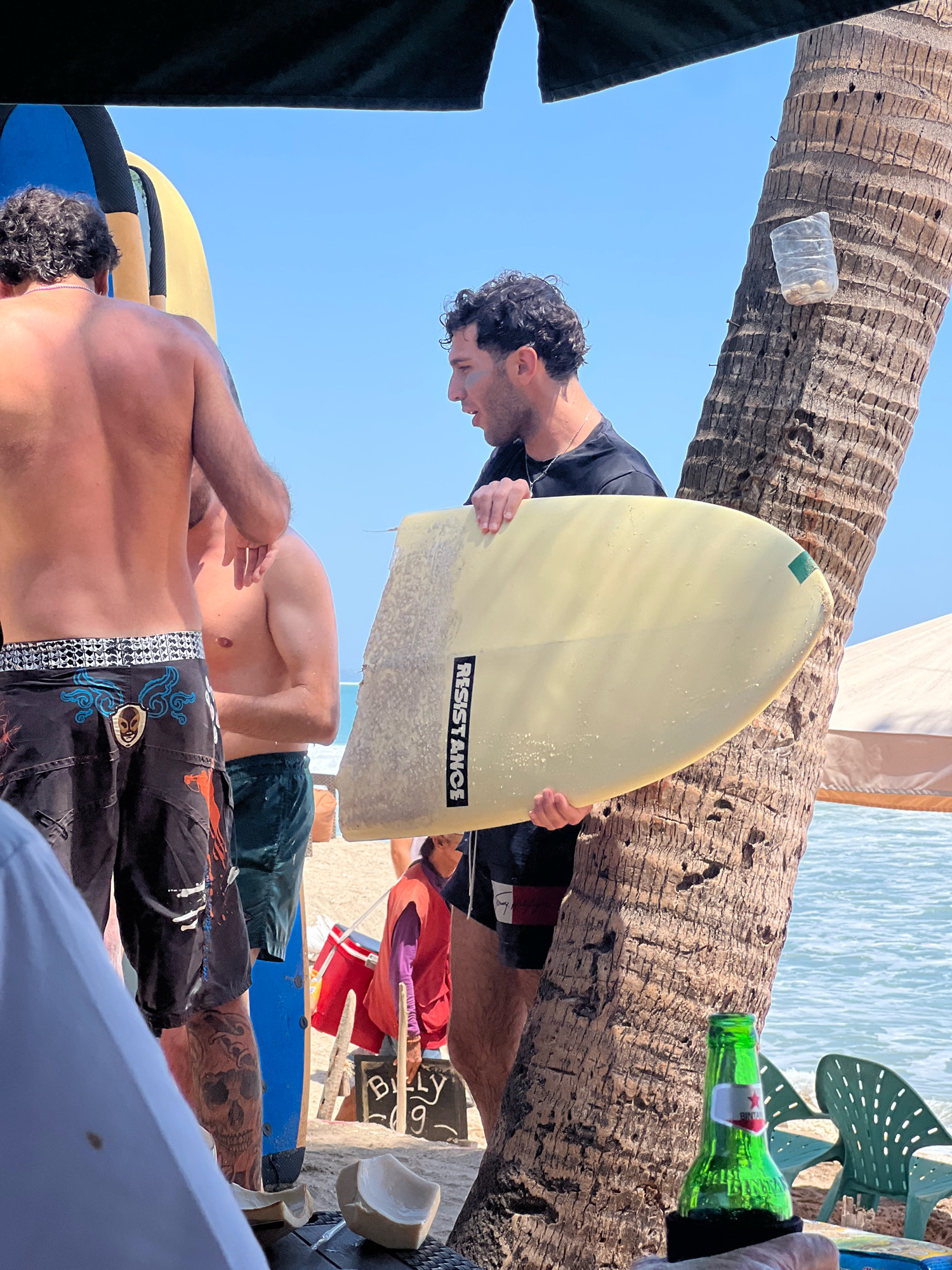 A close-up, eye-level shot captures a group of men on a sunny day at Kuta Beach, Bali, Indonesia, near a palm tree. In the foreground, the back of a man wearing black board shorts with intricate white and blue designs and a skull tattoo on his left calf is visible. To his right, another man, shirtless and with muscular arms, stands beside him. A third man, wearing a black t-shirt, is prominently featured holding a light-yellow surfboard with the word "RESISTANCE" printed vertically in black lettering on its side. The surfboard is angled towards the viewer, its rounded end visible. The palm tree, textured and weathered, serves as a backdrop, with a clear plastic bottle containing a few small objects affixed to its trunk. The ocean is visible in the distance, with gentle waves. A green beer bottle with a red and white label stands in a black koozie in the lower right corner, and a green plastic beach chair is positioned behind it. Other beachgoers and surfboards are partially visible in the background, as well as a tent-like structure. A sign with "BILLY 9" is visible on the sand. A broken coconut shell rests on a dark surface in the foreground. The scene suggests a relaxed atmosphere at a popular surf spot.