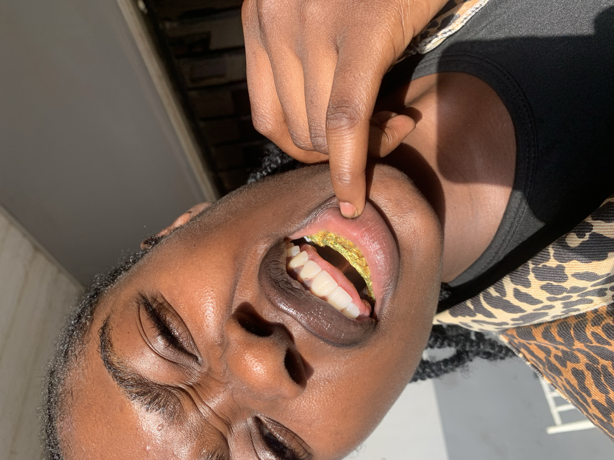 In the city of New Stadium, Nigeria, a high-angle, close-up photograph features an individual with dark skin. The subject's right eye is partially closed, displaying prominent facial lines around the eye and brow. The mouth is open, revealing white teeth and a crumpled sheet of reflective gold material positioned on the tongue. The lips are dark-toned and glossy. The subject's right hand is actively pulling down the left side of the lower lip with its fingers. Braided hair is visible on the left side of the frame. The individual wears a leopard-print outer garment over a dark-colored top. Strong, direct natural light illuminates the scene, creating distinct highlights on the skin. The background is out of focus, showing a grey overhead surface and a darker horizontal architectural feature in the upper right, along with a light-colored surface and a partial white object in the lower left.