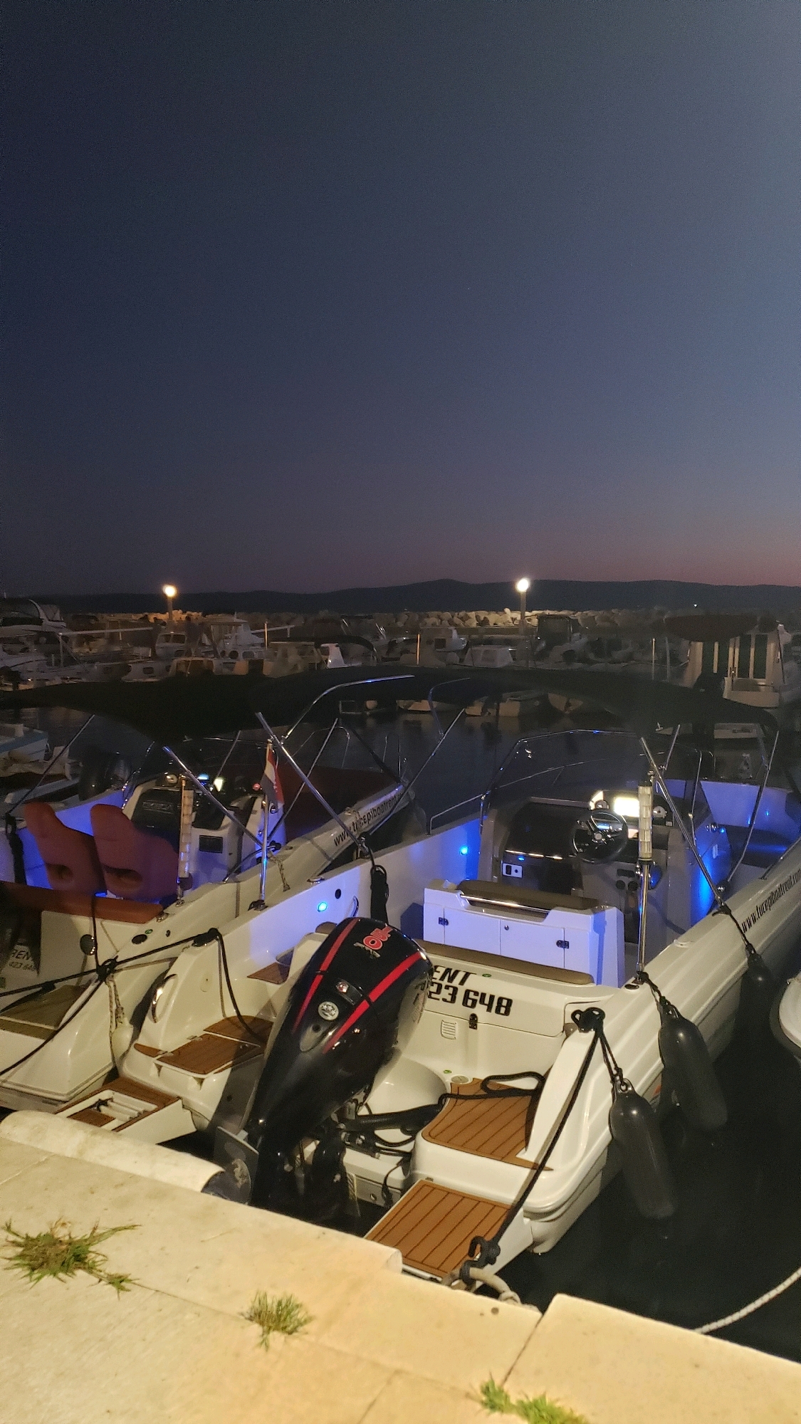 The image shows a marina in Općina Tučepi, Croatia, at dusk. Several boats are docked side-by-side, illuminated by subtle blue lights. The sky is a deep blue, transitioning to a lighter hue near the horizon. Distant hills are visible in the background, with lights from the town or other boats scattered across them. The foreground features a concrete walkway with patches of grass. One boat in the foreground has a black outboard motor with red accents. Another boat has red seats and a Danish flag. The overall scene is serene, suggesting a calm evening in the coastal town.