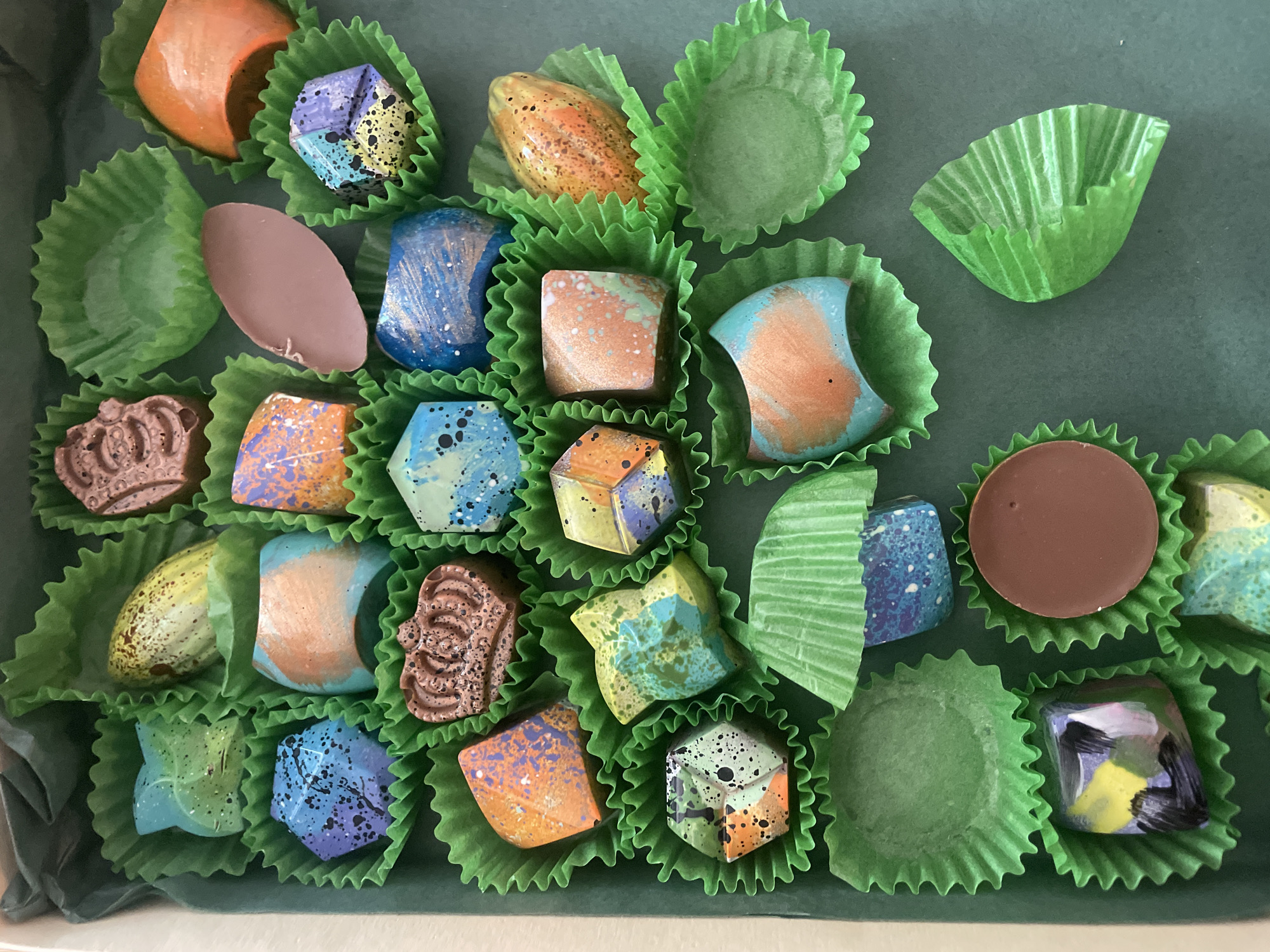 A top-down view shows a box of artisanal chocolates. The chocolates are of various shapes and colors, many with a speckled or painted appearance. They are arranged in green, fluted paper cups. The background appears to be a solid dark green. No people are visible. The image does not contain information that would allow me to determine the location of the scene within Sofia, Bulgaria, or to verify that it is in Sofia at all.