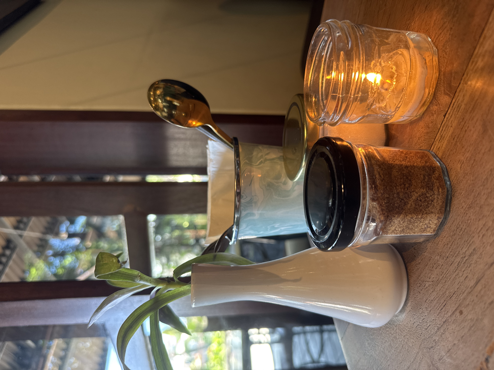 On a wooden surface, a table setting is arranged near a window that offers a view of greenery. The arrangement includes a small white vase with green leaves, a utensil holder with a golden spoon, a spice jar with a black lid, and a lit candle in a clear glass jar. The scene is likely photographed at a restaurant or cafe in North Kuta, Indonesia, indicated by the tropical flora visible through the window.