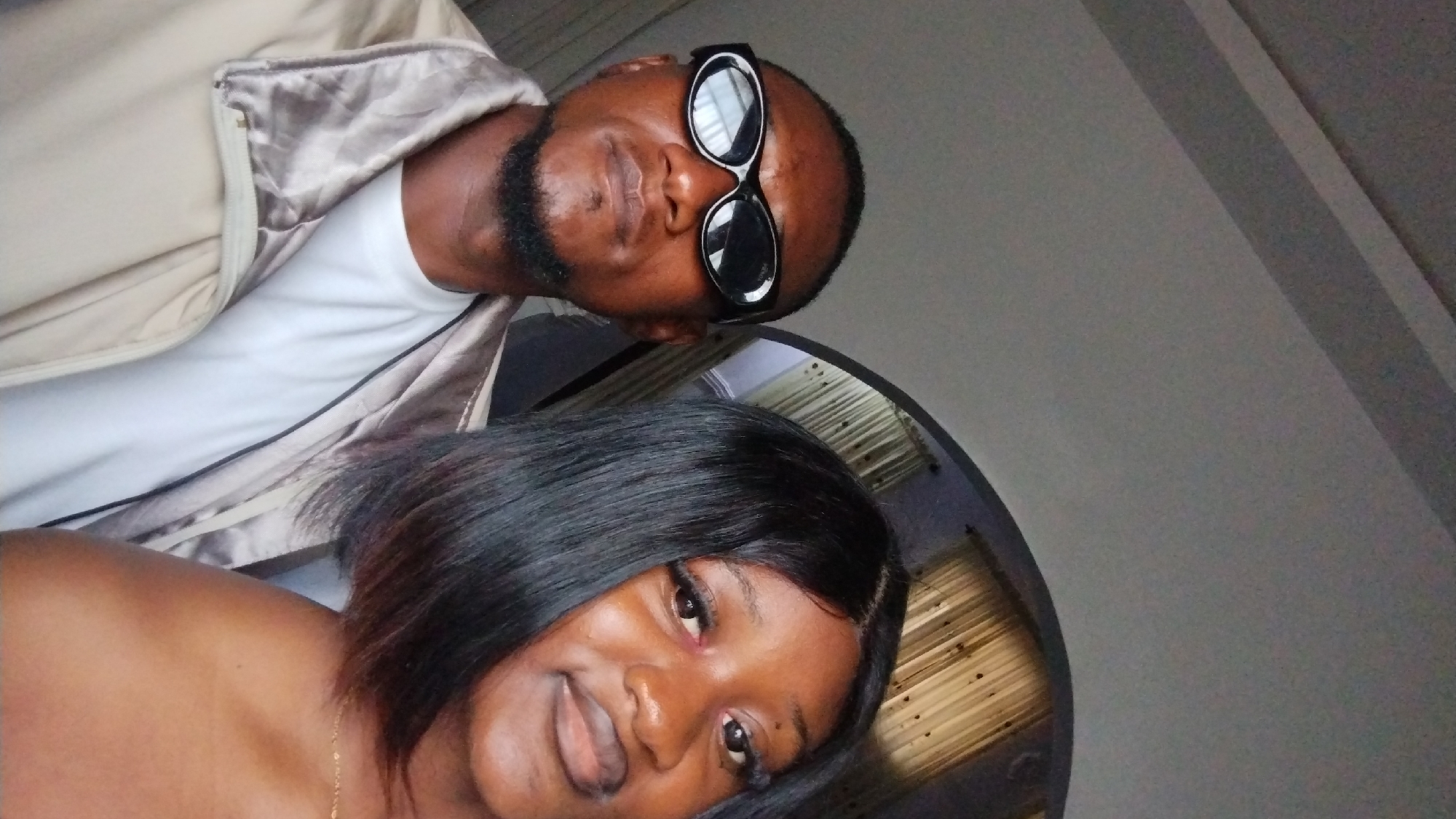 A man wearing black sunglasses and a white T-shirt under a light-colored jacket is posing for a selfie with a woman in what appears to be an indoor setting. The location is possibly in the city of Jos, Nigeria, given the provided context. The woman has short, black hair and is smiling. The background is a neutral-colored wall.