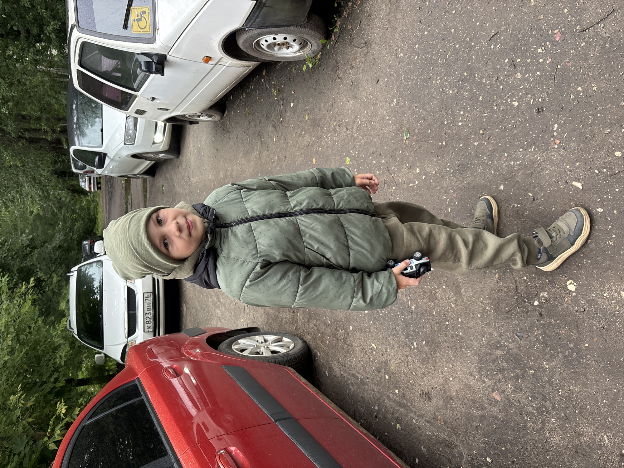 A young boy stands on a dirt road, looking at the camera. He is wearing a green hooded jacket and a matching beanie. He is holding a toy car in his right hand. Behind him, several cars are parked, including a red sedan on the left and white vehicles on the right. Trees and foliage line the background. The scene appears to be in an outdoor setting, possibly a parking lot in Rusanovo, Russia, given the license plate on the white car. The overall lighting suggests an overcast day.