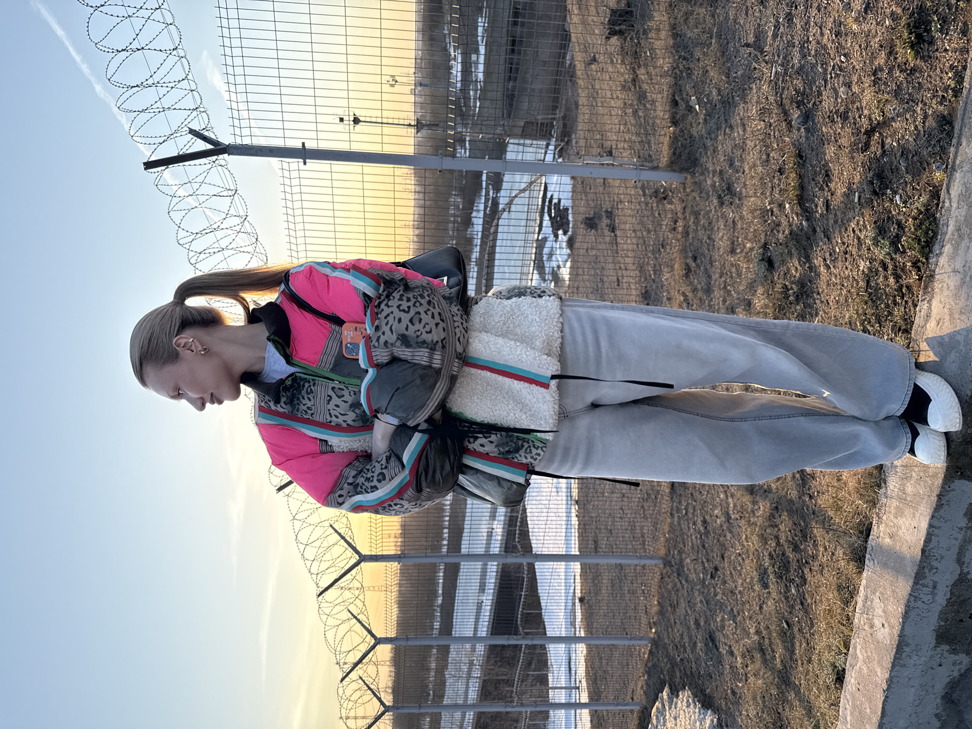 A young woman with blonde hair pulled back in a ponytail stands near a fence topped with barbed wire. She wears light-colored jeans, white sneakers with black trim, and a colorful jacket in shades of pink, blue, and gray. She has a backpack draped over her shoulder. The area behind the fence appears to be a field with patches of snow. The photo was taken in the city of Chaikovsky, Russia. The background suggests an open area with industrial structures. The sun appears to be setting or rising, based on the light's angle and color. A concrete barrier is in the foreground.