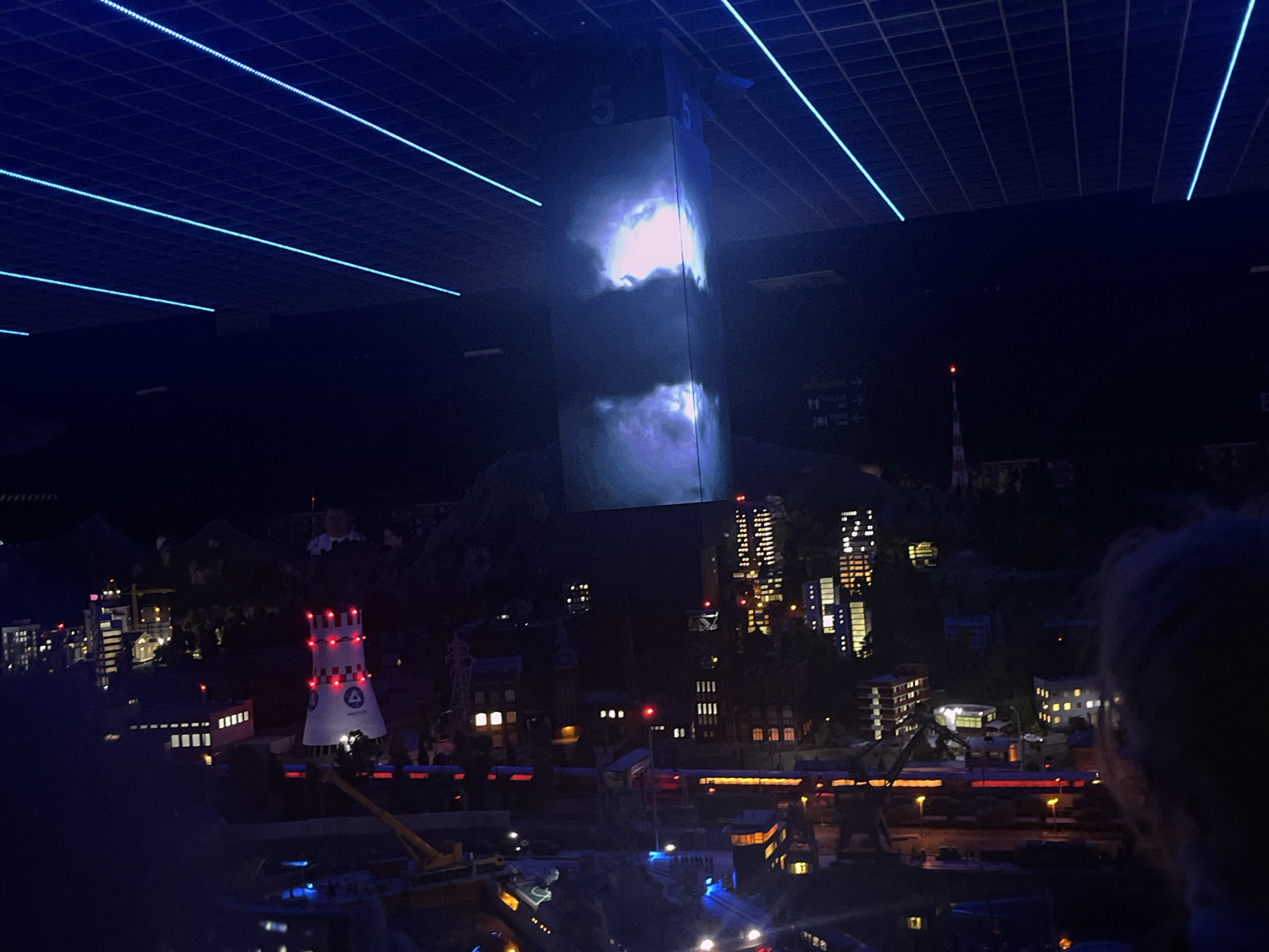 The image depicts a miniature model of a city, likely Saint Petersburg, Russia, viewed from a slightly elevated perspective at night. The city model features numerous buildings of varying heights and architectural styles, many of which are illuminated with internal lights, creating a glowing effect against the dark backdrop. Prominent in the foreground is a distinctive white tower with red accents and lights, possibly representing a landmark or industrial structure. Other notable elements include a construction crane, trains on tracks, and what appears to be a harbor or waterfront area.
Above the city, a large, rectangular screen is projected onto a vertical surface, displaying a cloudy, atmospheric image with bright, ethereal light emanating from within the clouds. The ceiling above the model is a dark grid structure with thin, bright blue LED lights running in parallel lines, creating a futuristic or theatrical ambiance. The presence of a person's head and shoulder on the right side of the frame indicates that the viewer is likely at an indoor exhibition or attraction. The overall scene suggests a nighttime view of a city, enhanced by artificial lighting and projected imagery.