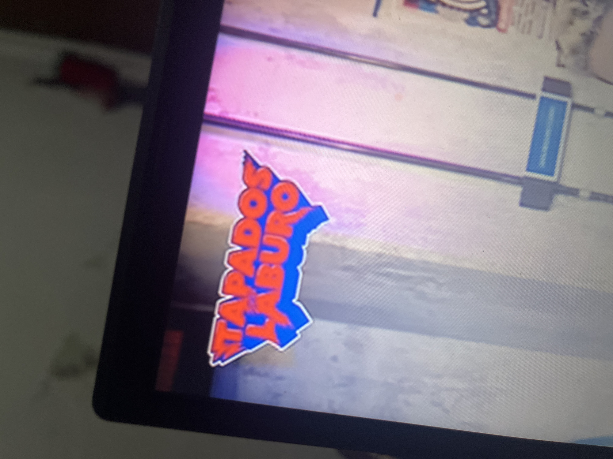 The image shows a close-up, slightly blurry view of a computer screen displaying a logo with the text "TAPADOS DE LABURO". The logo is shaped like a map of Venezuela with flames and a blue outline. The background on the screen appears to be a bright, somewhat abstract image with vertical lines and color gradients. The surrounding environment is dark, with the screen being the primary light source. There are no discernible people or specific locations within Santa Ana, Venezuela visible in the image. The focus is entirely on the screen content.