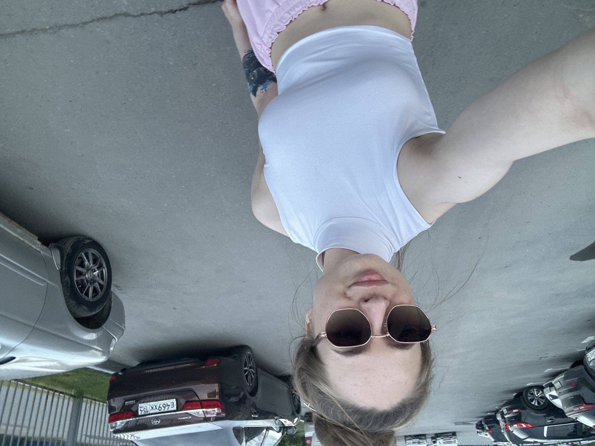 The image is a selfie taken by a young woman outdoors. She is wearing a white tank top and pink shorts, and sunglasses. She has a tattoo on her left arm. The background shows a parking lot with several cars, including a silver car and a dark-colored sedan. The ground is asphalt, and there is a metal fence visible in the distance. The image is oriented upside down. The time of day and weather are not discernible from the image.