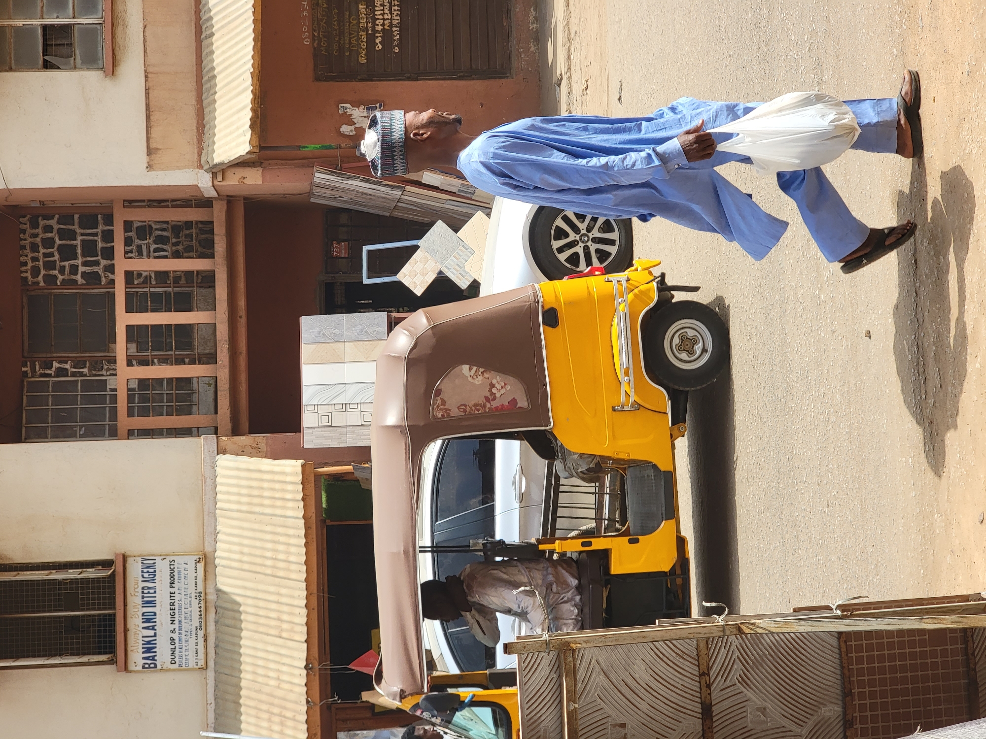 A man in a light blue traditional dress and a patterned hat is walking on a sunny street in Maiburiji, Nigeria. He is carrying a white plastic bag in his right hand. Behind him is a parked yellow and brown auto rickshaw. An individual is sitting inside the rickshaw. A white car is parked directly behind the rickshaw. The buildings on the left side of the street include a sign for "BANKLAND INTER AGENCY" which lists them as a dealer in Dunlop & Nigerite products. Additional tiling can be seen leaning against the side of the building above the auto rickshaw. A sign with handwritten phone numbers is visible on the upper-right building.
