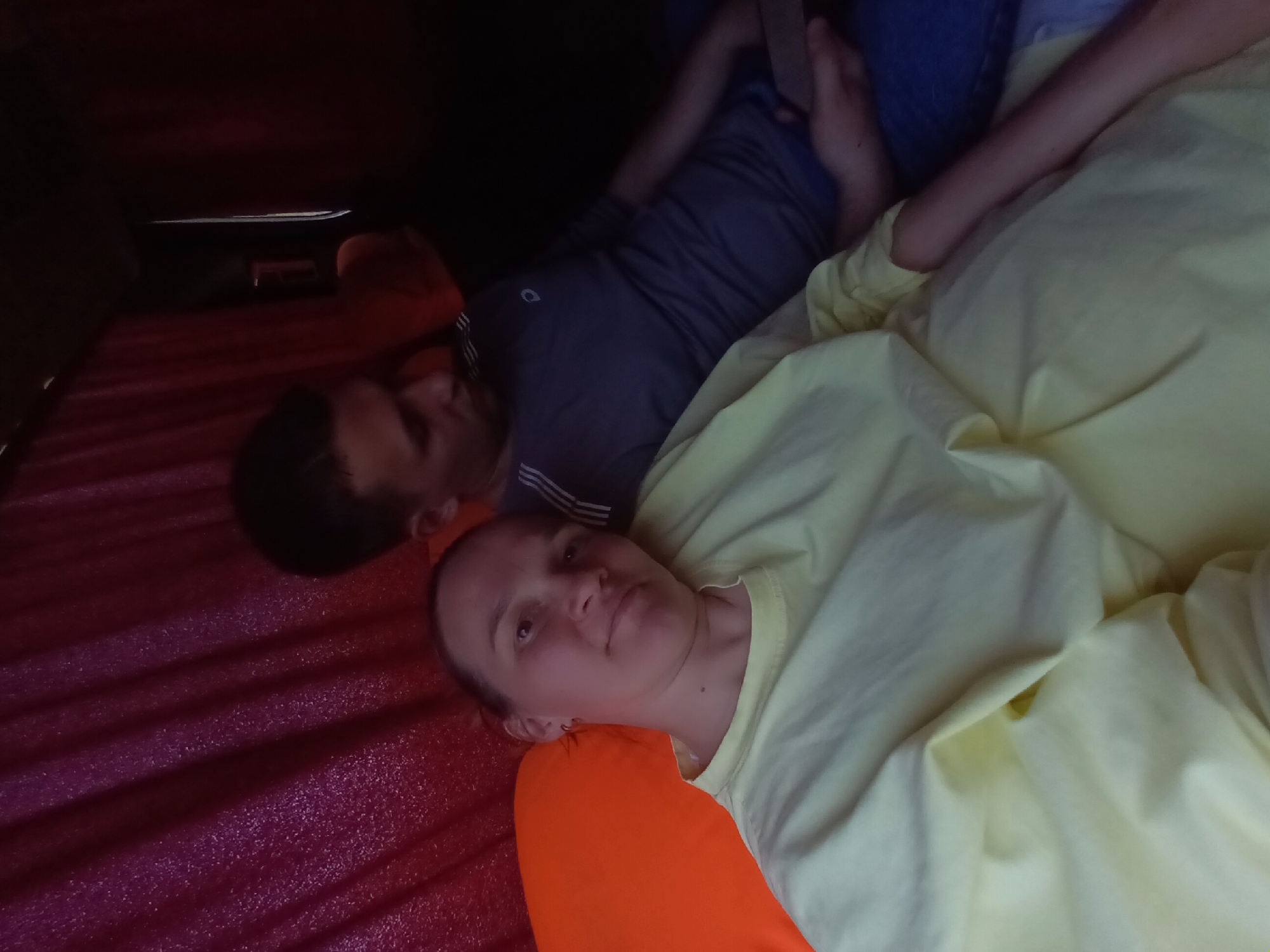 A couple is lying down inside what appears to be a tent or enclosed space. The woman is in the foreground, wearing a yellow shirt and resting her head on an orange pillow. Behind her, a man lies down wearing a grey shirt, also with an orange pillow. The surface beneath them is a maroon color and appears to be a mattress or sleeping pad with a ribbed texture. The lighting is dim, suggesting they are indoors or in a sheltered area. The location is not evident from the image.