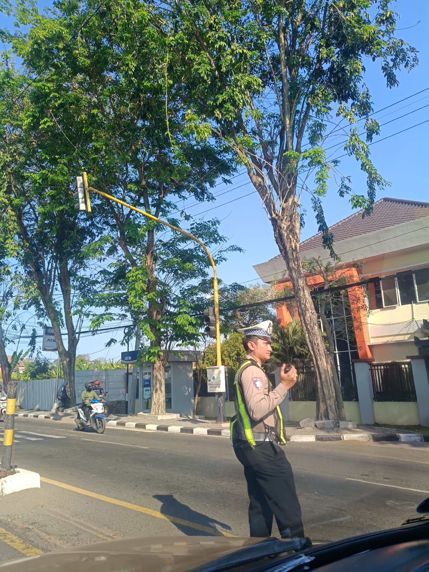 The image captures a street scene in Klangonan, Indonesia, under a clear blue sky. In the foreground, a traffic police officer stands on the road, wearing a white hat, a light brown uniform with a reflective vest, and dark pants. He appears to be holding a communication device in his right hand. The officer is positioned on the right side of the frame, facing left.
The road is paved with asphalt and marked with yellow and white lines. A yellow traffic light pole with a traffic signal is visible to the left of the officer. Several trees with dense green foliage line the street, with their branches reaching towards the sky. In the background, a building with an orange and white facade and a tiled roof is visible.
Further down the road, a motorcycle with two riders is seen moving away from the viewer. Another motorcycle is parked on the side of the road. There are also signs and a fence in the background, suggesting a built-up area. The overall impression is a busy street intersection in an Indonesian town.