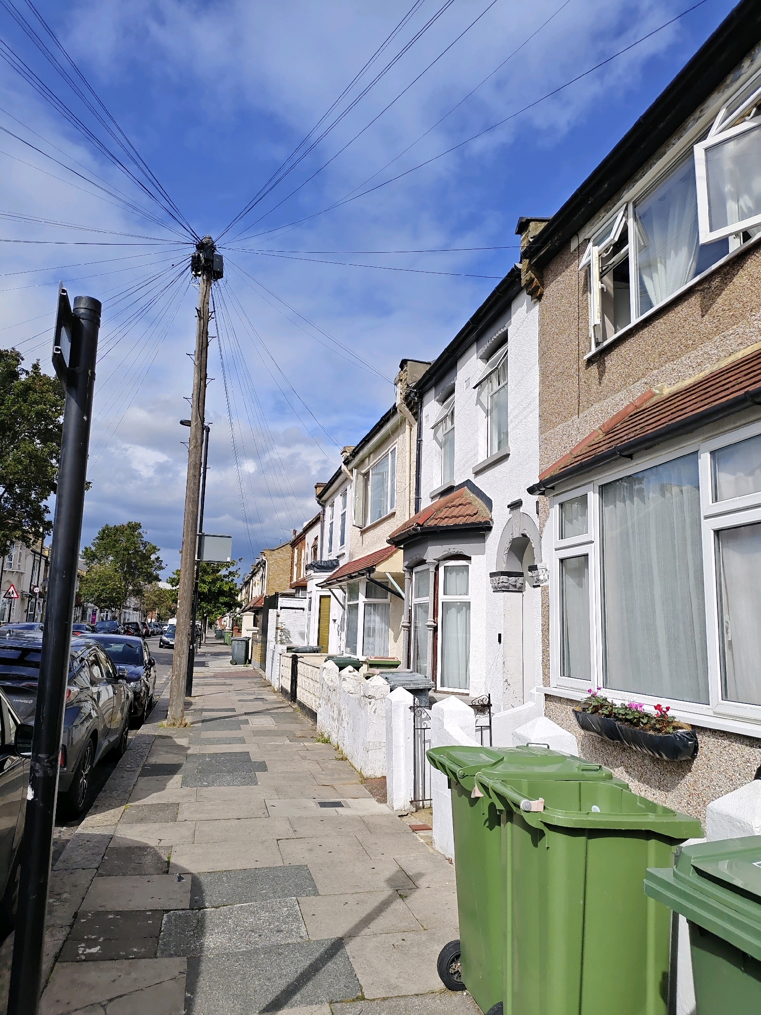 This is an eye-level, outdoor, full shot of a street in London, United Kingdom, on a partly cloudy day. The perspective is from the sidewalk looking down the street, towards a line of terraced houses on the right. A prominent wooden utility pole with a multitude of wires radiating from its top stands on the left side of the street, with a black pole in front of it. Cars are parked along the left side of the street, and garbage bins are positioned on the sidewalk in front of the houses on the right. The houses themselves are modest, with a mix of pebbledash and white render exteriors, and feature bay windows and red-tiled roofs. The sidewalk is paved with concrete slabs of varying sizes and colors.