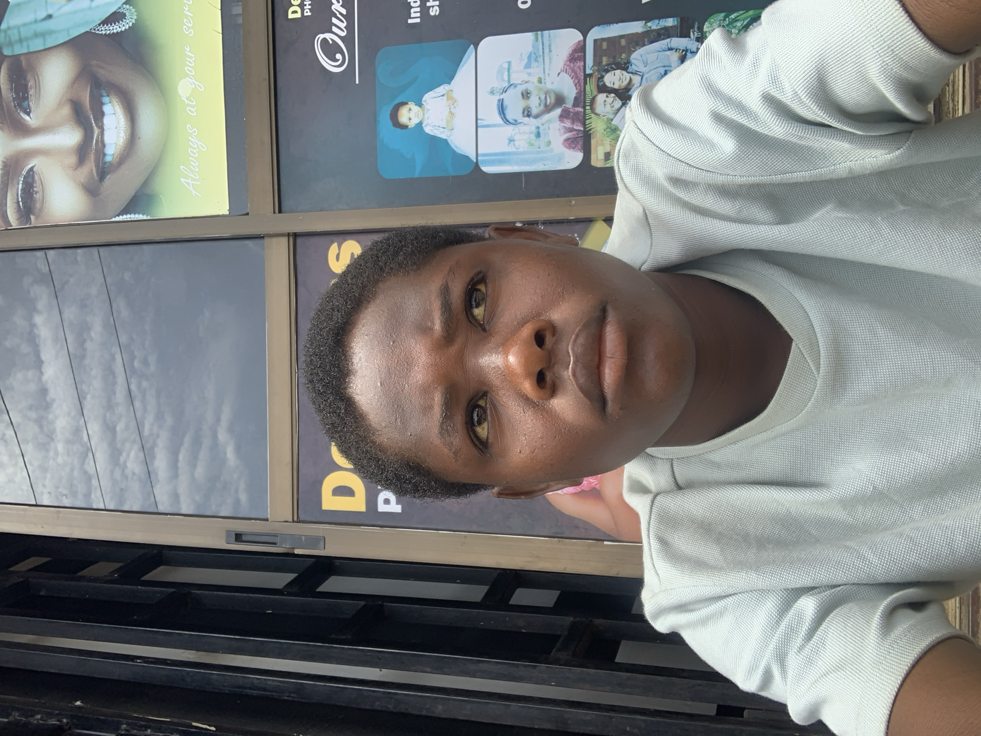 A close-up, high-angle photograph of a dark-skinned person with short, dark hair wearing a light-colored, long-sleeved shirt. The person is looking at the camera with a neutral expression. Behind the person are posters and reflections, suggesting the photo was taken indoors near a window with promotional material displayed. The bottom of the frame includes part of a metal structure, possibly a gate or railing.
The posters visible include images of people, and text like "Always at your service" and "Our". Based on the poster content, the location appears to be near a photography studio or related service. The location is specified as Barakin Akawo, Nigeria.