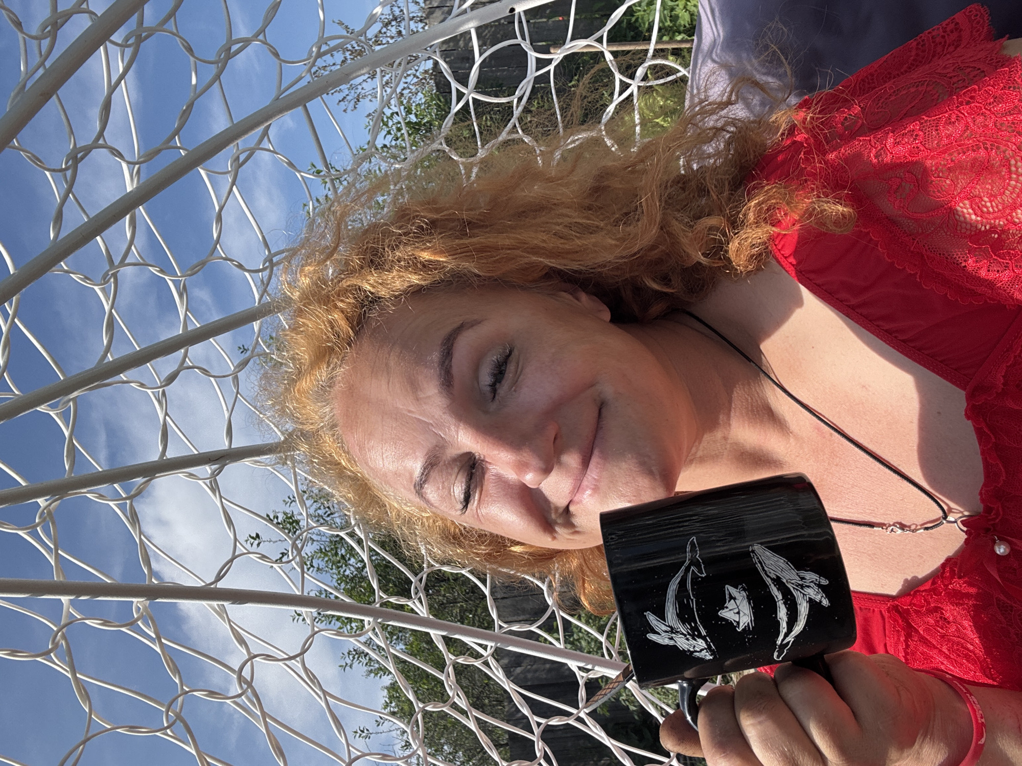 A woman with curly red hair is holding a black mug with white illustrations of whales and a paper boat. She is wearing a red lace top and a necklace with a pendant. The woman is smiling with her eyes closed and appears to be in an outdoor setting, possibly a garden, with a white wire structure behind her and trees and a blue sky visible. The image is taken on a sunny day. The location is Chaikovsky, Russia.