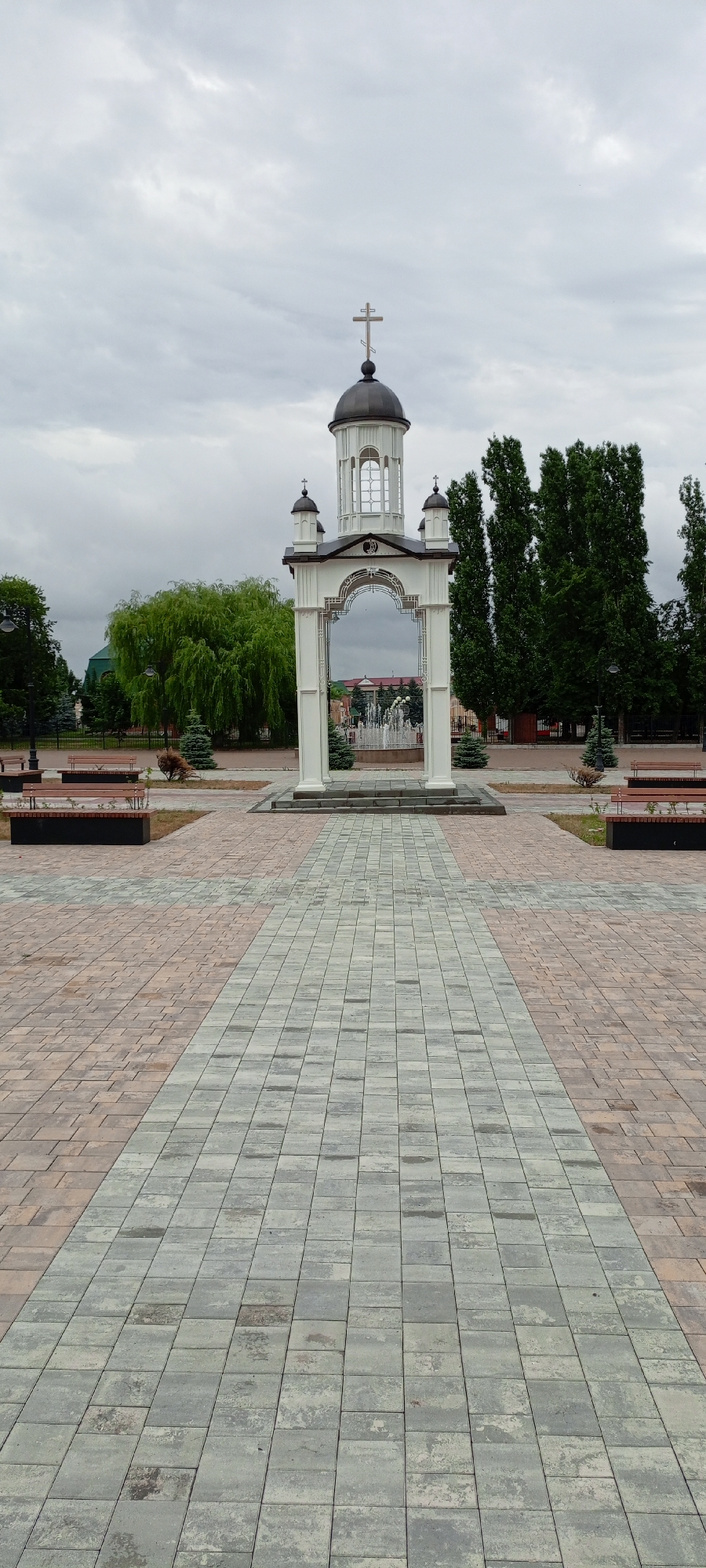 The image depicts a paved public square in Kirsanov, Russia. In the center stands a white ornate structure resembling an archway topped with a dome and a cross, suggesting a religious or memorial significance. Through the archway, a fountain with multiple jets of water is visible. The square is paved with two-toned bricks, arranged in a patterned walkway leading towards the central structure. To the sides of the walkway, there are benches and some greenery, including trees and bushes. The sky is overcast, casting a soft, diffused light over the scene. The overall impression is that of a serene and well-maintained public space.