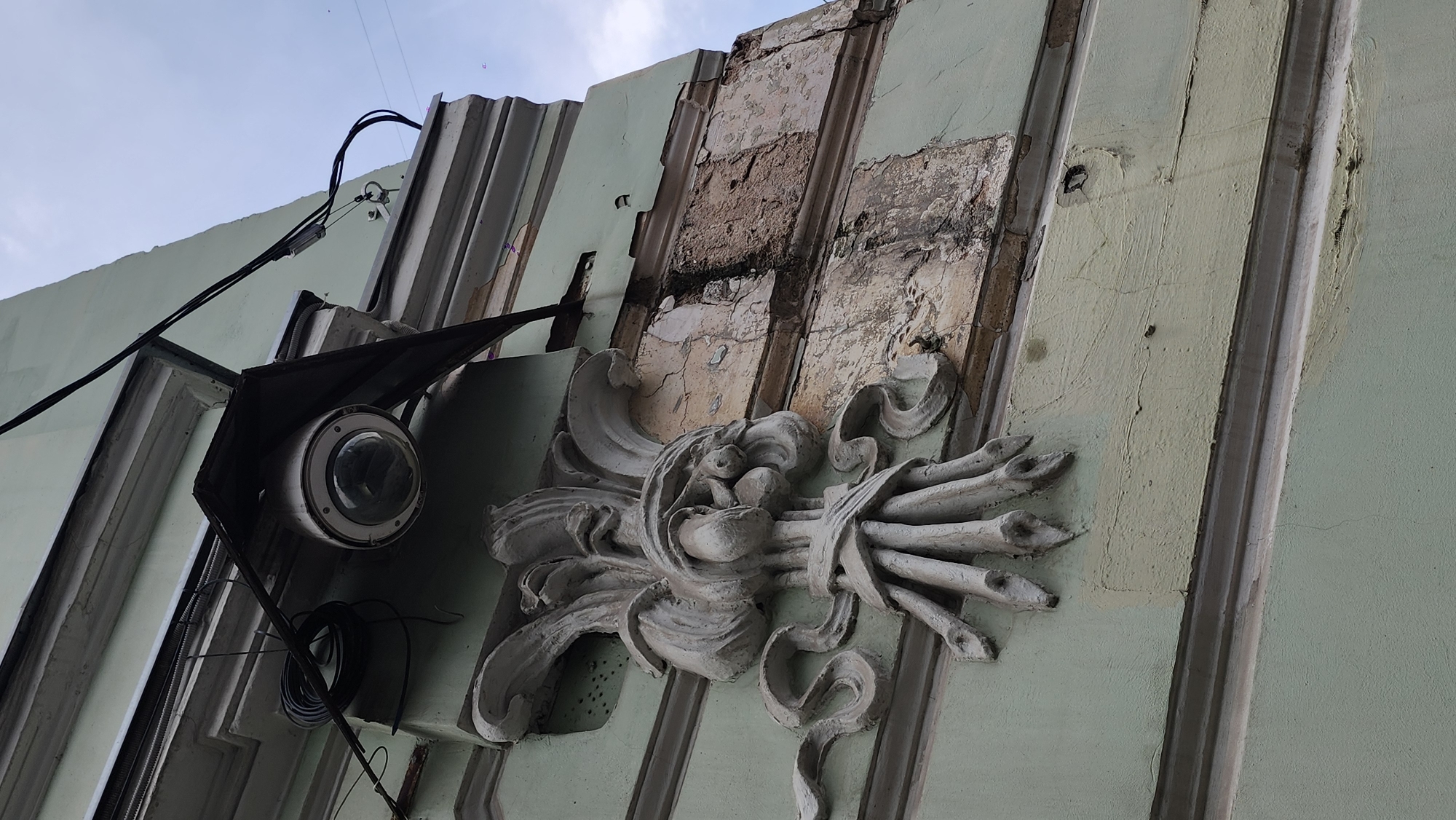 A low-angle shot depicts the facade of a weathered building. On the left is a security camera mounted on a metal structure. Various wires connect to it, running along the building's exterior. Adjacent to the camera is a decorative, sculpted ornament affixed to the wall. The wall itself shows signs of decay, with crumbling sections revealing the inner layers of the building's construction. The building's color is light green.
The picture was taken in Moscow, Russia.