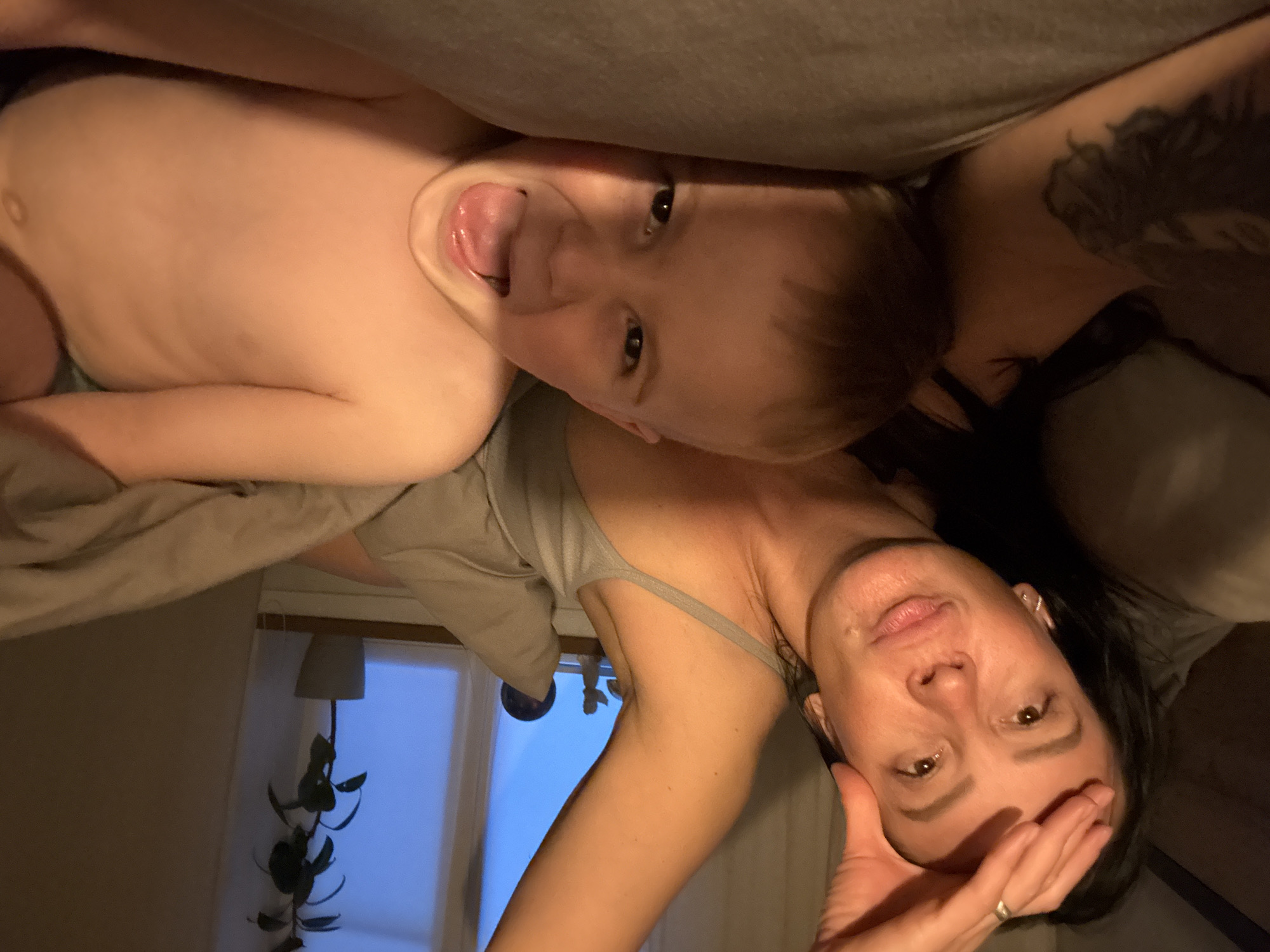 A woman and a young child are posing for a selfie. The woman, with dark hair, is wearing a grey tank top. Her right hand is near her forehead and she has a ring on her finger. She has a tattoo on her upper right chest. The child, shirtless, is looking directly at the camera and sticking his tongue out. The child is resting on the woman. They are in a bedroom, with a window in the background. A lamp and a plant are visible near the window. The location is Chaikovsky, Russia.