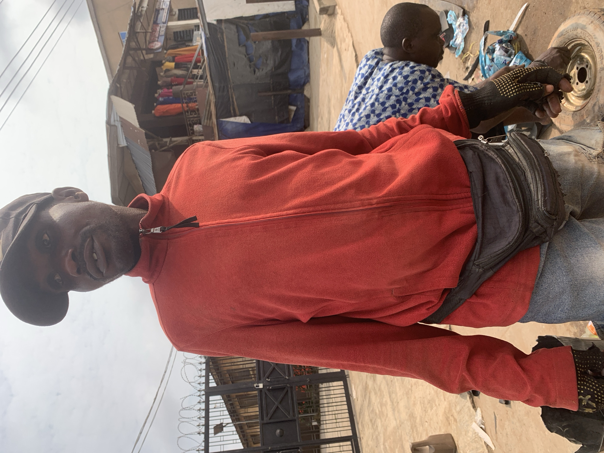 A man in a red zip-up jacket and a baseball cap stands facing the camera, his face partially obscured by the cap. He wears a black fanny pack around his waist and denim jeans. His right hand, wearing a black and yellow-dotted glove, is raised and holds a piece of what appears to be a tire. In the background, another person is bent over a car wheel on the ground. The environment appears to be an outdoor urban setting in Shagamu, Nigeria, with a fenced gate and buildings visible behind the man. The ground is dirt and debris.