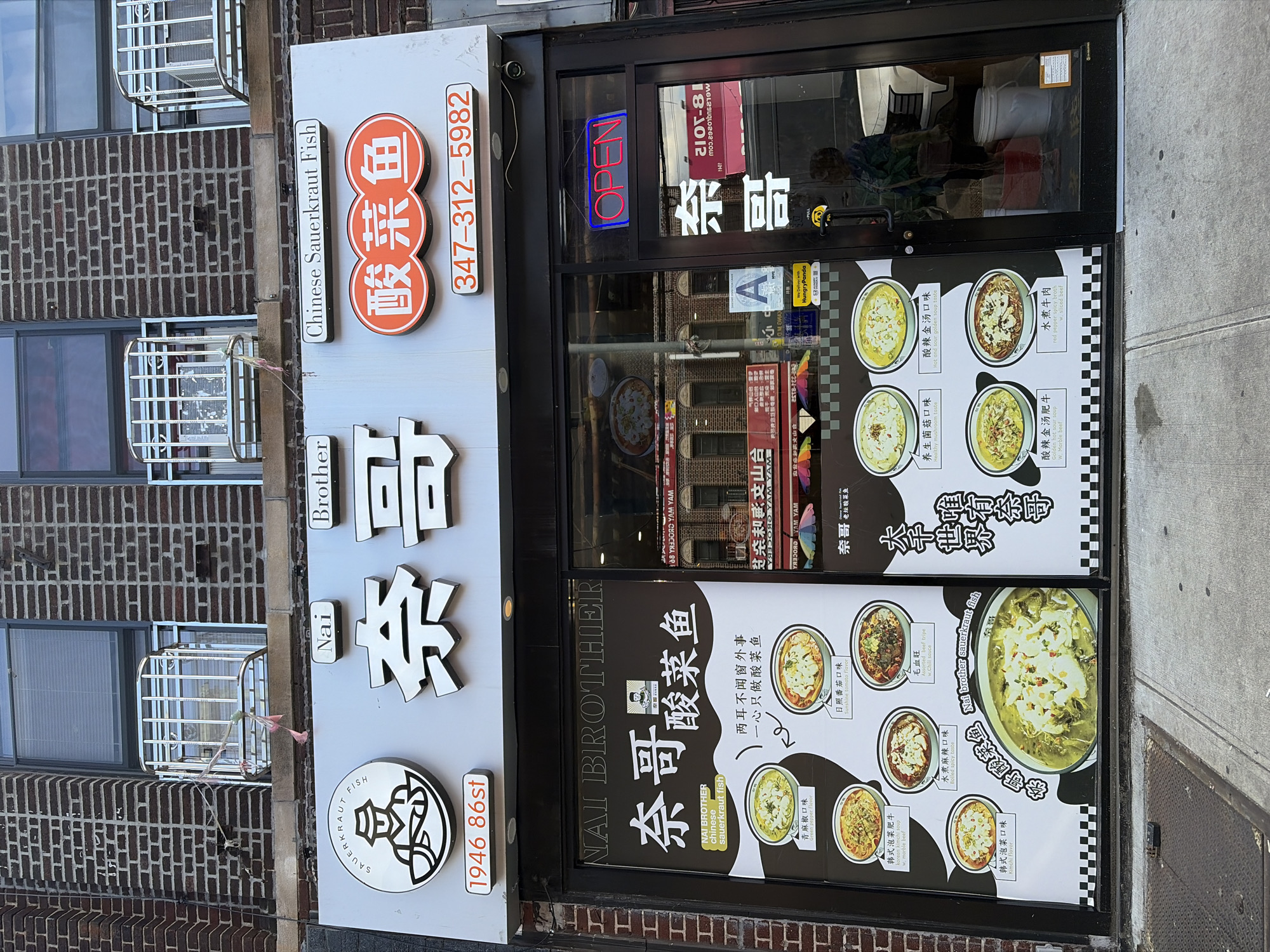 The image shows the exterior of a restaurant called "Nai Brother Chinese Sauerkraut Fish" located at 1946 86th Street in Brooklyn, New York. The restaurant has a prominent sign with its name in English and Chinese characters, along with a phone number. The storefront features a large window displaying a menu with pictures of various dishes, all appearing to be soups or stews with fish and vegetables. Some of the dishes are labeled in English with flavors such as "Green pepper flavor," "Sunshine tomato flavor," and "Kimchi flavor." An "OPEN" sign is illuminated in red and blue neon lights in the window. The building is made of brick, and there are windows with metal security bars on the upper floors. The street in front of the restaurant is paved with concrete and has a metal grate. A person can be seen reflected in the glass of the door.