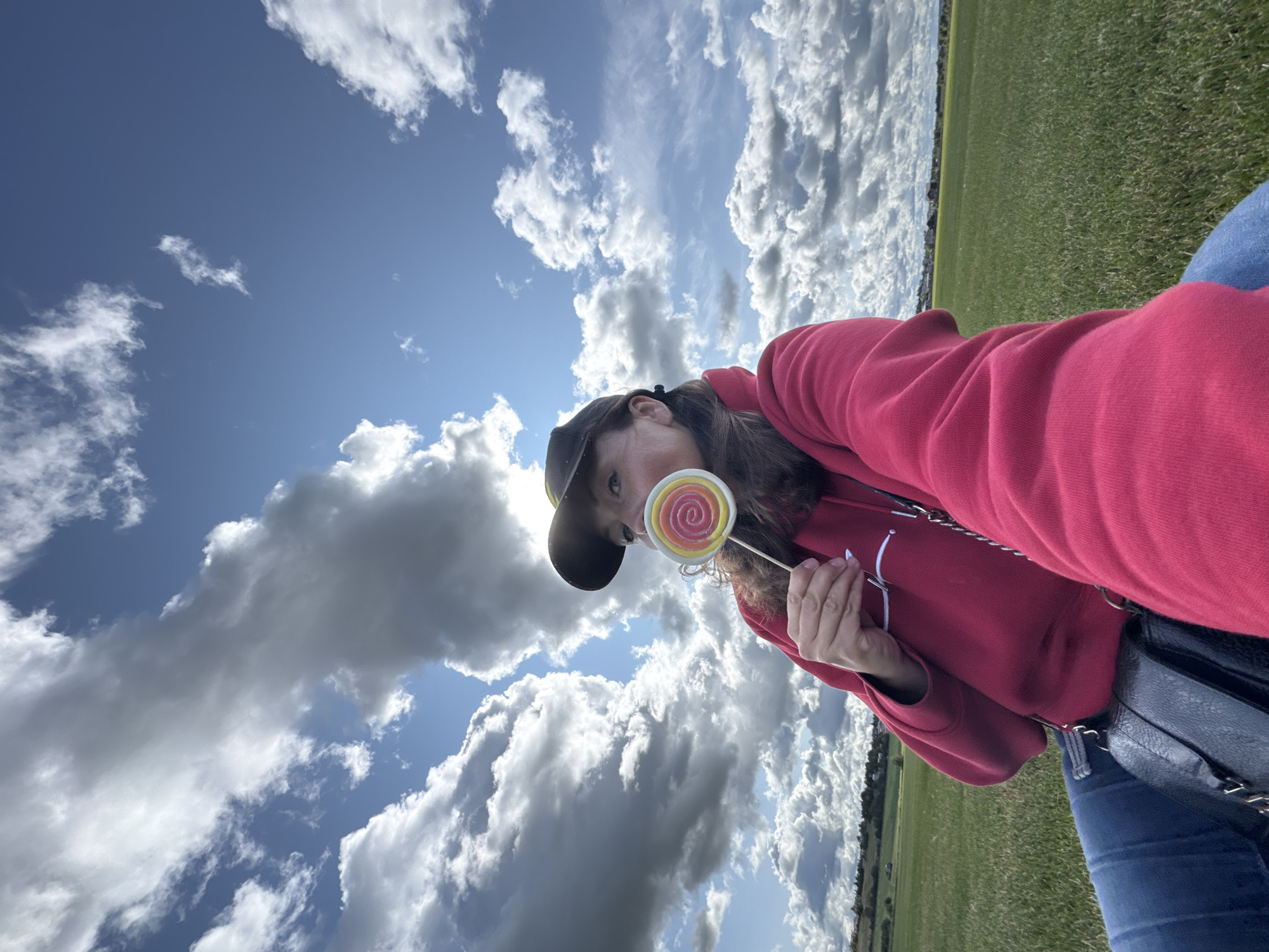 A low-angle, selfie-style shot captures a person wearing a red hoodie and blue jeans, sitting in a grassy field under a partly cloudy sky. The person is holding a swirled lollipop up to their mouth, partially obscuring their face. A black purse with a chain strap is visible at their hip. In the background, the field extends to a horizon line with distant trees and buildings, suggesting a rural or suburban setting. The sky is a mix of blue and dramatic, white and gray clouds. The image does not contain any information that would allow for its location to be identified as Innolovo, Russia.