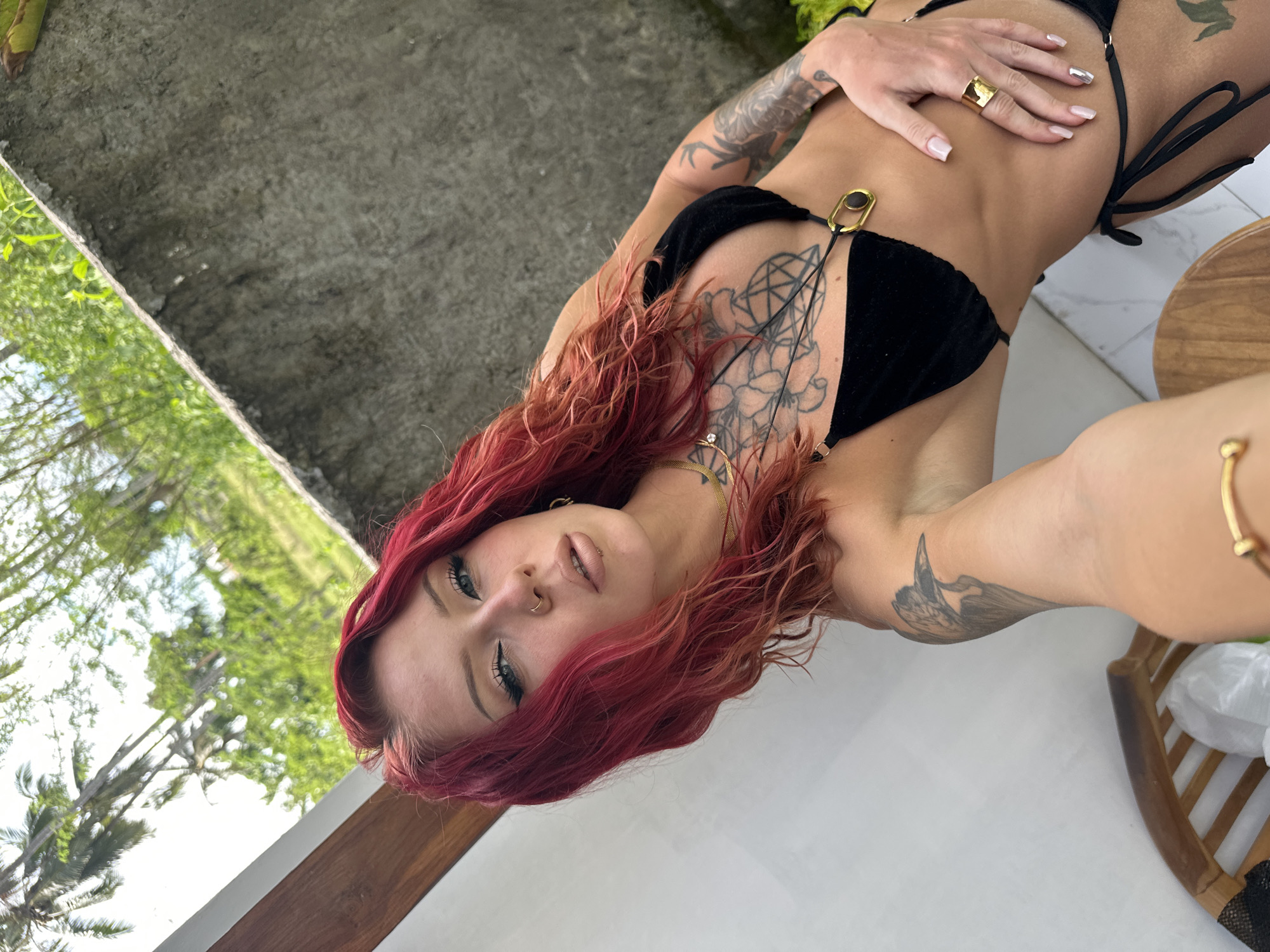 A woman with red hair poses for a selfie. She is wearing a black bikini and several pieces of jewelry. The background includes a concrete surface, greenery, and trees. The location appears to be in Nusa Penida, Indonesia.