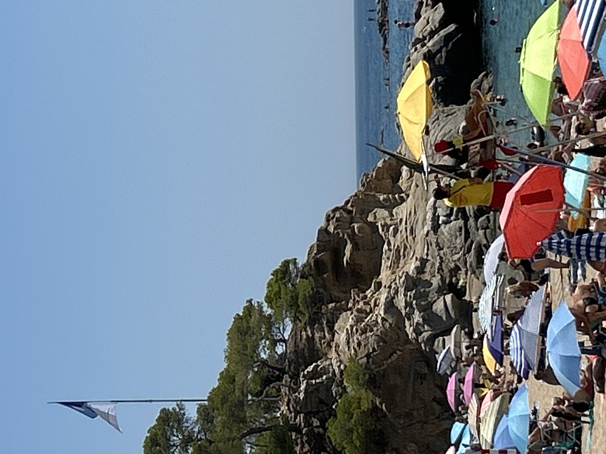 The image depicts a crowded beach scene in Castell d'Aro, Platja d'Aro i s'Agaró, Spain. Numerous beachgoers are spread across the sand, many seated under colorful umbrellas of various shades, including blue, pink, yellow, and red. A lifeguard in a yellow shirt and red shorts sits on an elevated chair, observing the water. The beach is backed by rocky cliffs dotted with green trees. The sea, a deep blue, is visible in the background with several people swimming or wading. A flag flies from a pole on the cliff, and a bright yellow umbrella is prominent in the foreground.