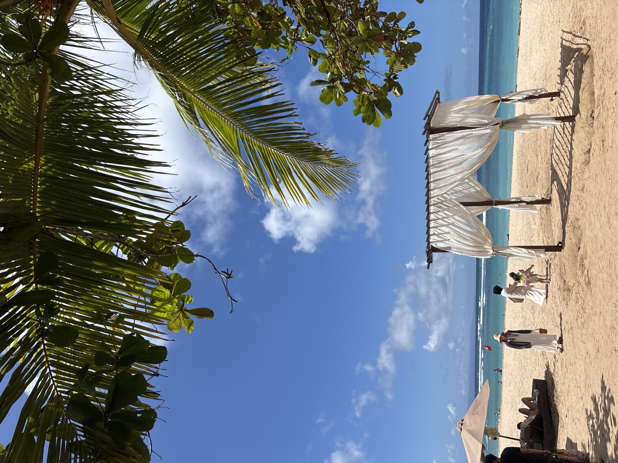Captured from a low angle in Kuta Selatan, Indonesia, the photograph depicts a beach scene under a vibrant blue sky dotted with cumulus clouds. A palm frond and other foliage frame the upper portion of the shot. On the sandy beach, a white-draped wedding gazebo stands, indicating a potential ceremony or event. Several individuals are visible in the scene, including people interacting near the gazebo and others lounging under an umbrella in the foreground. The turquoise ocean forms the backdrop, with gentle waves lapping the shore.