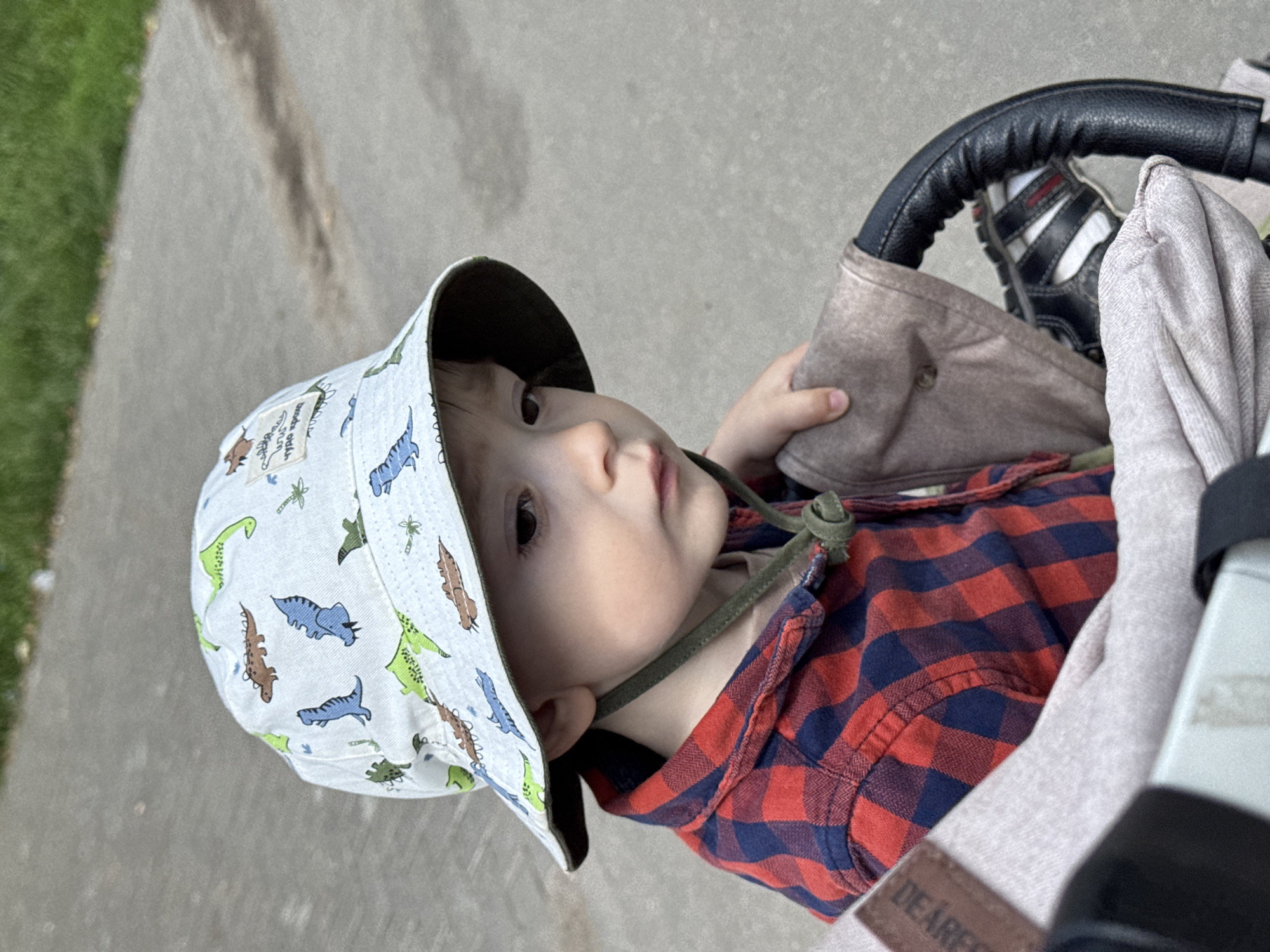 The image shows a close-up of a young child sitting in a stroller. The child is wearing a light-colored bucket hat with a dinosaur pattern and a red and blue plaid shirt. The child's face is turned to the side, and they appear to be looking down. The stroller has a brown fabric seat and a black leather handle. The background is a paved surface with some green grass visible. The scene is likely outdoors. It is not possible to determine the specific location within Izhevsk, Russia from the image alone.