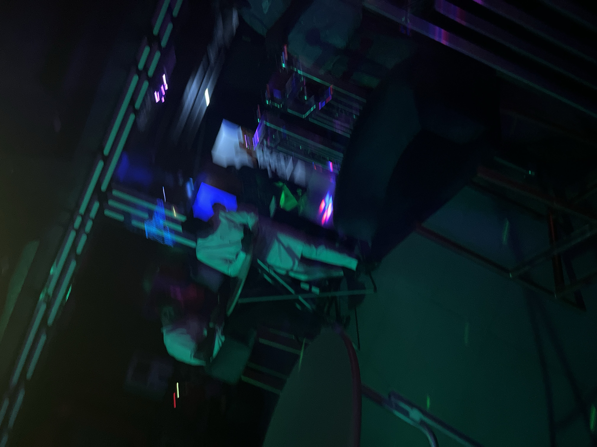 In an indoor venue, bathed in a predominantly green and blue neon light, two individuals are captured in motion. One, dressed in white, stands near a metal table and chair setup, seemingly engaged with an object in their hands. Another figure, also in white attire, is partially visible to their left, facing away from the camera. The background is characterized by the dim glow of neon lights lining what appears to be a ceiling or wall structure, and hints of other patrons and seating arrangements are discernible in the blurred periphery. The overall impression is that of a lively, dimly lit environment, possibly a club or lounge.