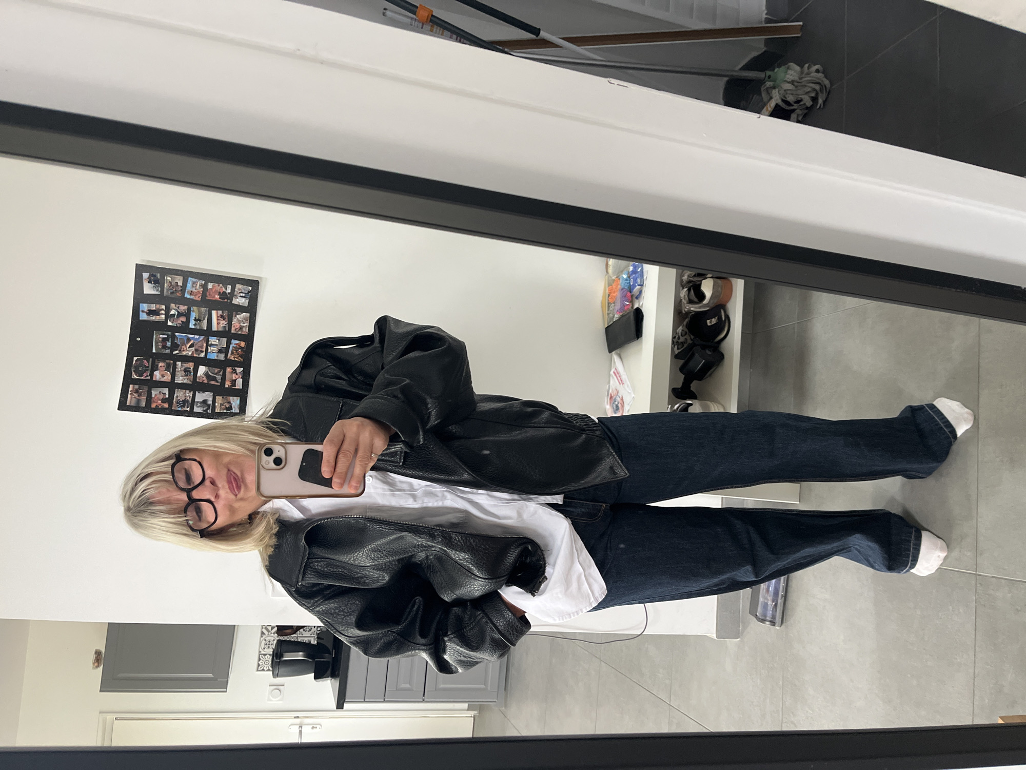 A blonde woman wearing a black leather jacket, a white shirt, dark-colored jeans, and white socks, with glasses, takes a mirror selfie with a light-colored phone. The reflection shows a kitchen with grey cabinets and tiled floors. A collage of photos is visible on the wall behind the woman. A mop leans against the wall. The location is Chambœuf, France, based on available information.