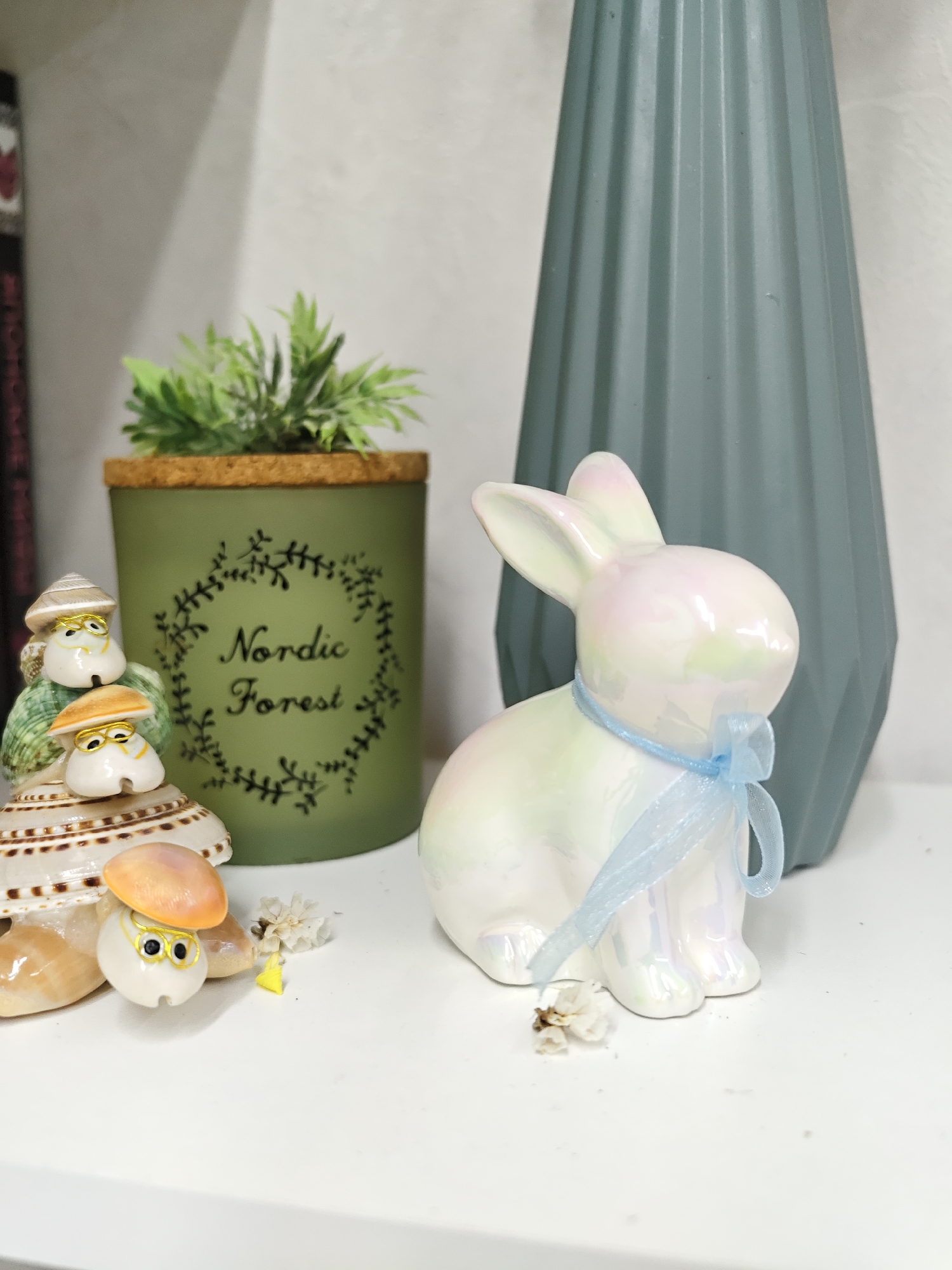 The image presents a still life arrangement featuring decorative objects on a white surface. From left to right, a shell figurine with multiple characters, a green candle holder labeled "Nordic Forest" with artificial green foliage on top, a pearlescent ceramic rabbit with a light blue ribbon tied around its neck, and a blue-gray vase with a fluted design. The objects are placed close together, suggesting a deliberate composition. The background is a plain wall. It is not possible to ascertain the location as Chaikovsky, Russia, from the given image.
