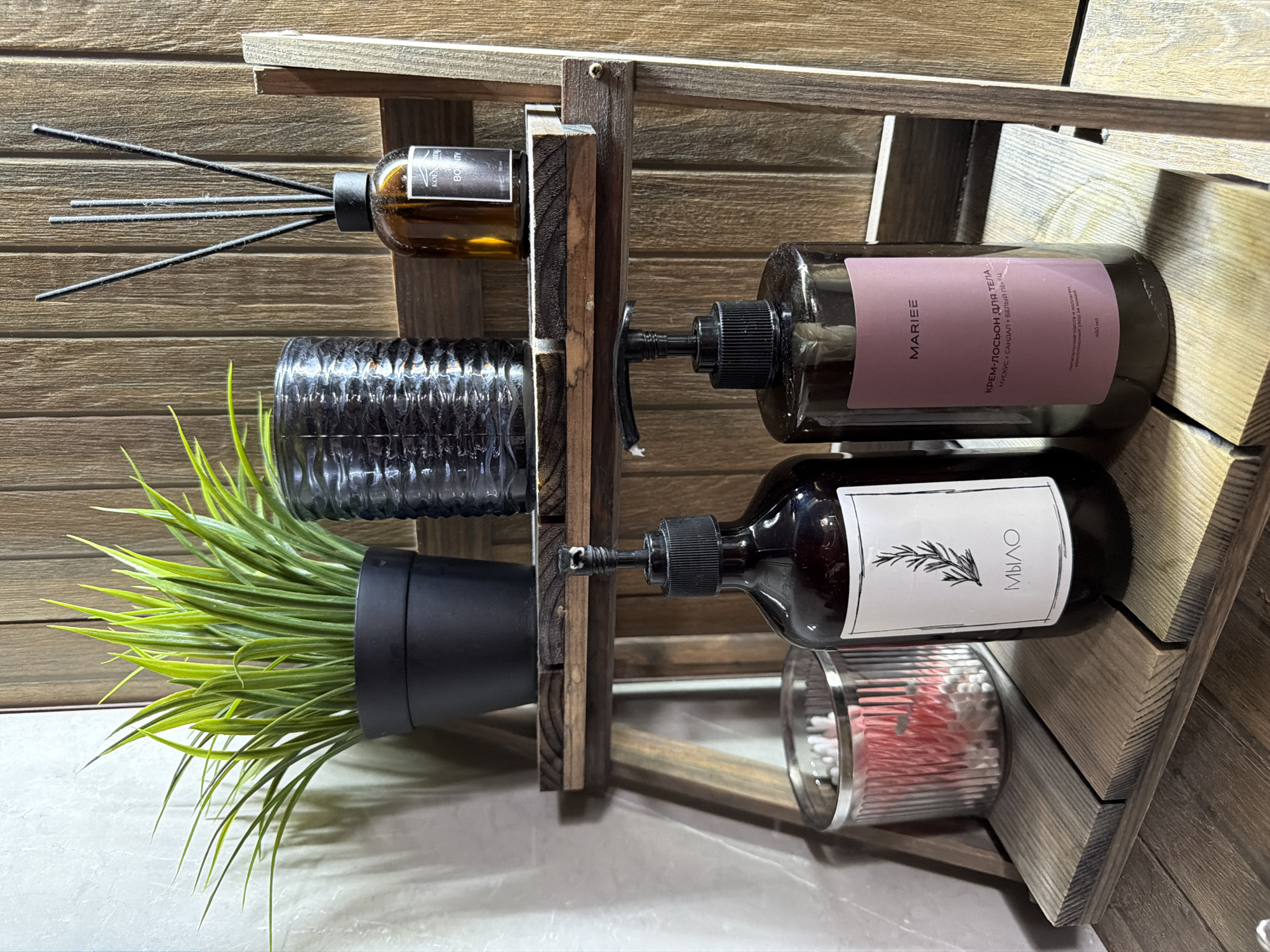 The image depicts a two-tiered wooden shelf unit, possibly for bathroom or vanity storage, against a textured wooden plank wall. On the top shelf, a small potted green artificial plant sits to the left, next to a dark, ribbed glass container and a brown glass diffuser bottle with black reeds. On the bottom shelf, a clear glass container filled with cotton swabs and some pink items is positioned to the left. To its right, there are two dark pump-top bottles. The left bottle has a white label with a sprig illustration and the Cyrillic word "МЫЛО" (soap). The right bottle has a pink label with the text "MARIEE" and further Cyrillic text indicating a body cream-lotion. The overall scene is well-lit and suggests a domestic setting. There are no people present in the image, and no actions are being performed. The environment is static and organized. There is no information in the image to indicate the location in the city of Семилуки, Russia.